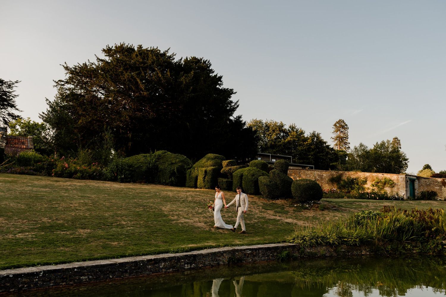 Wedding couple strolling on grassy hillside near a pond at sunset with trees in distance.
