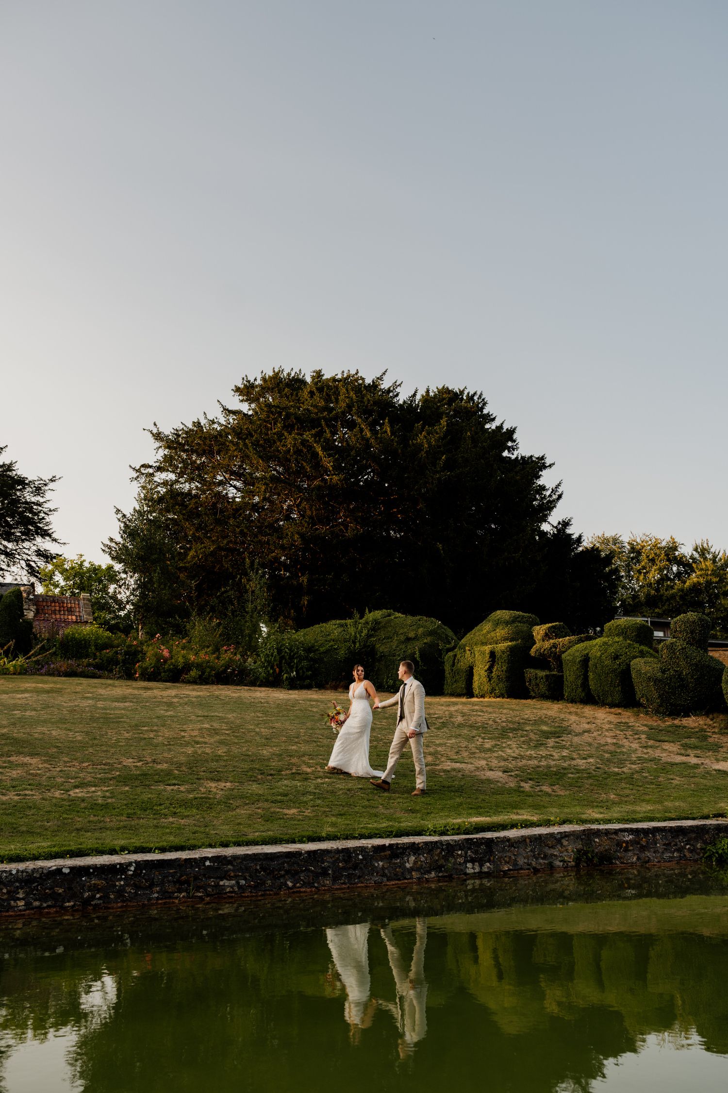 Couple in wedding attire walking along a reflective pond during golden hour with trees in background.