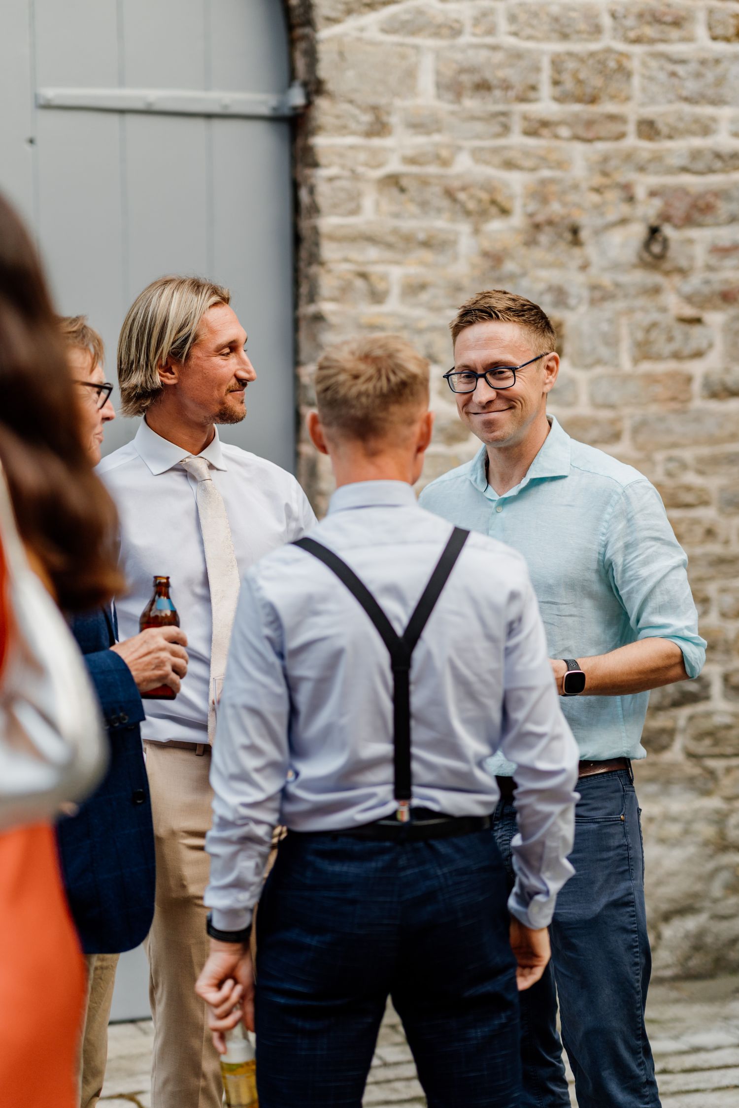 Wedding party guests in suspenders and light blue shirts gather outside a stone building.