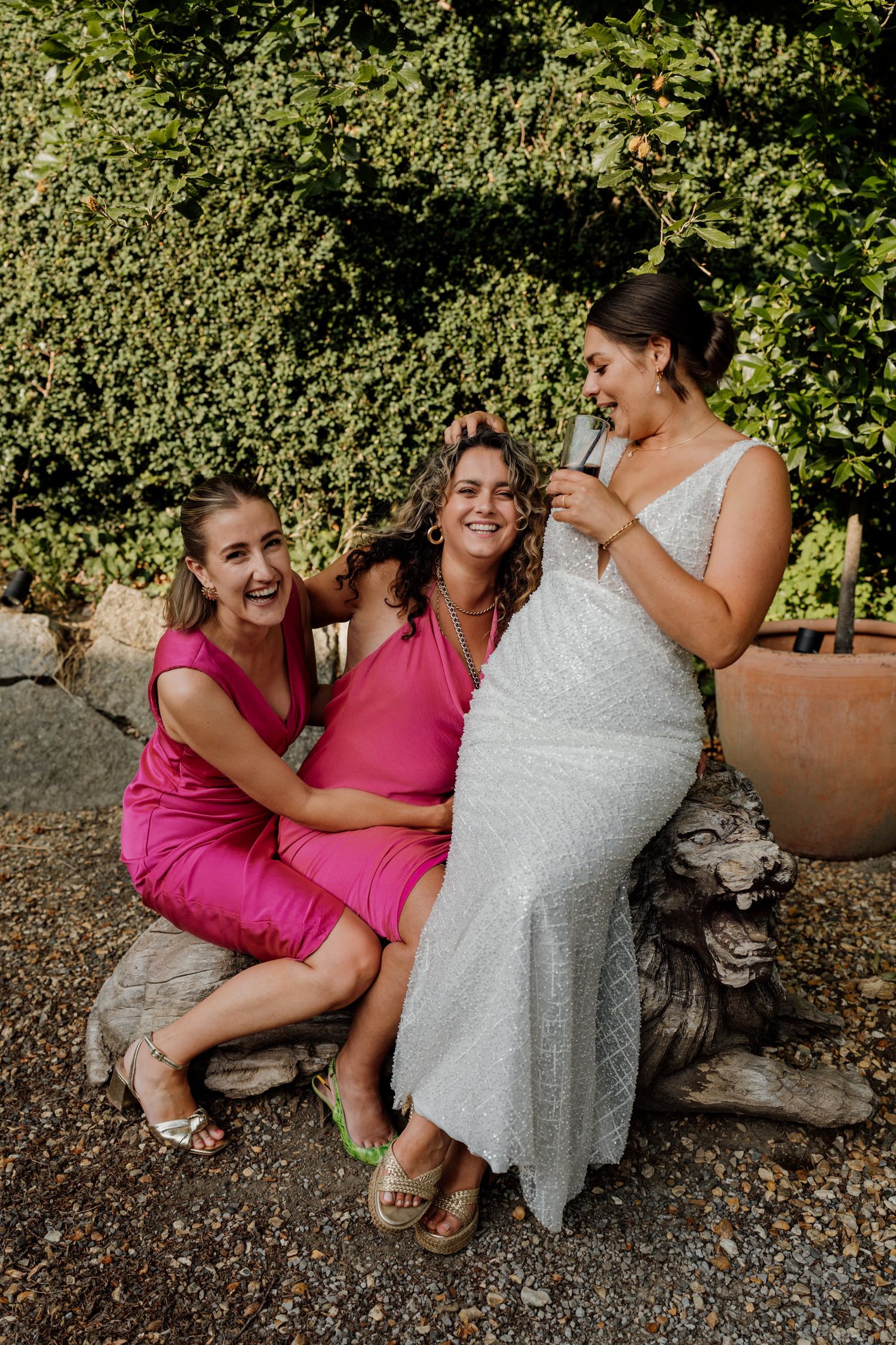 Friends in pink dresses and a white wedding gown share laughter while sitting on stone steps in an outdoor garden setting.