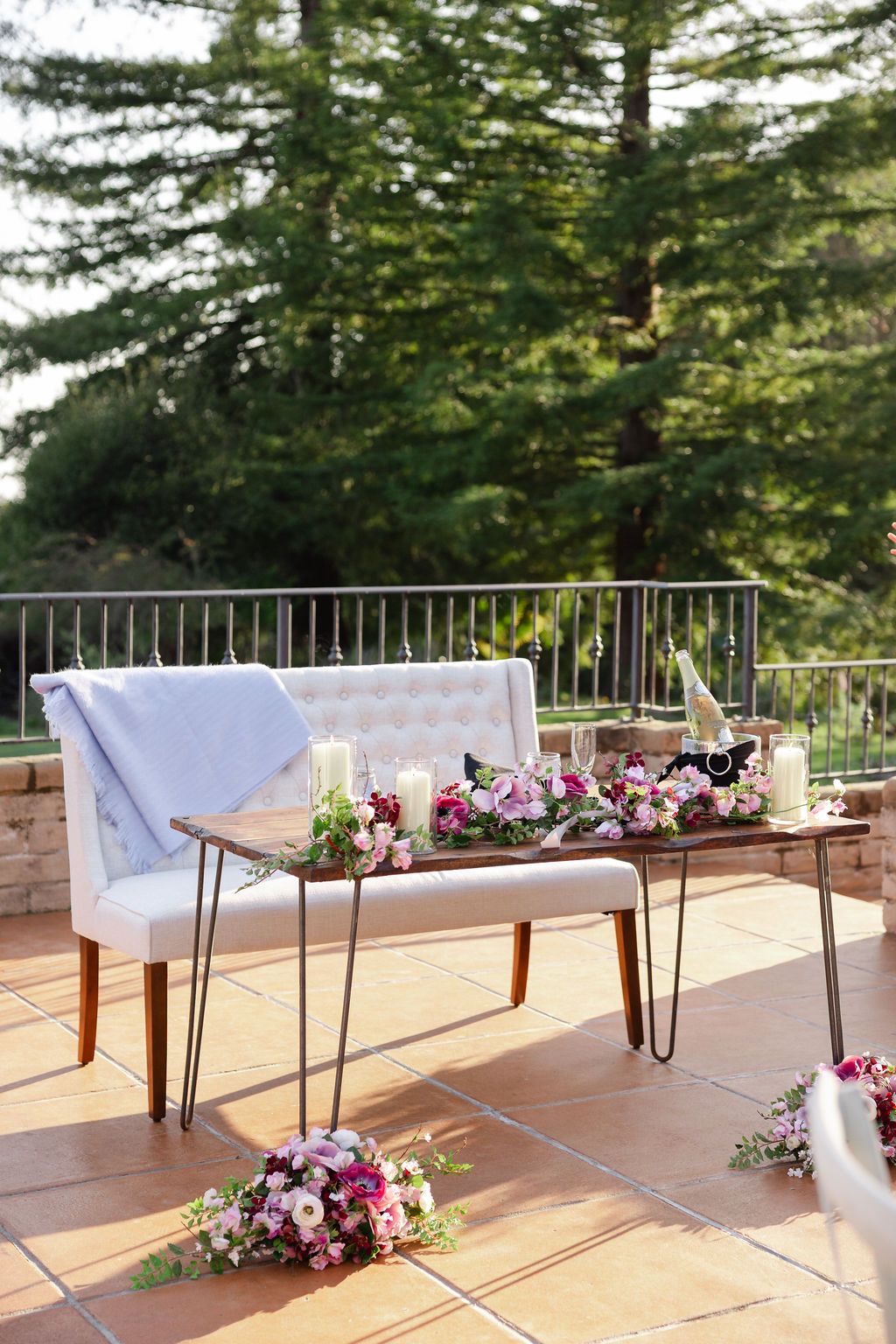 Simple white table settings with elegant pink floral arrangements on a sunny outdoor patio deck surrounded by evergreen trees.