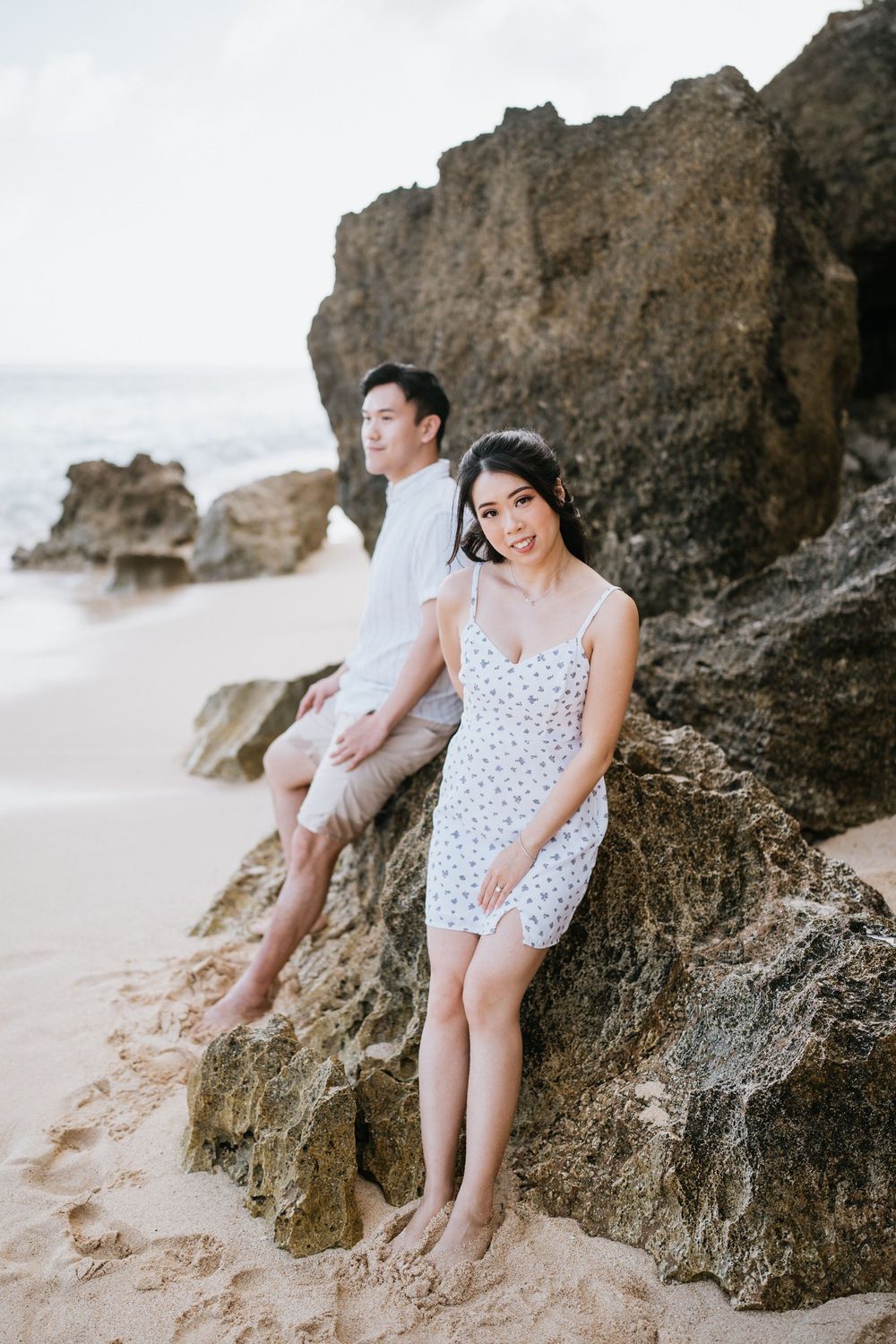 Romantic beach photoshoot with couple sitting on coastal rocks near ocean waves.