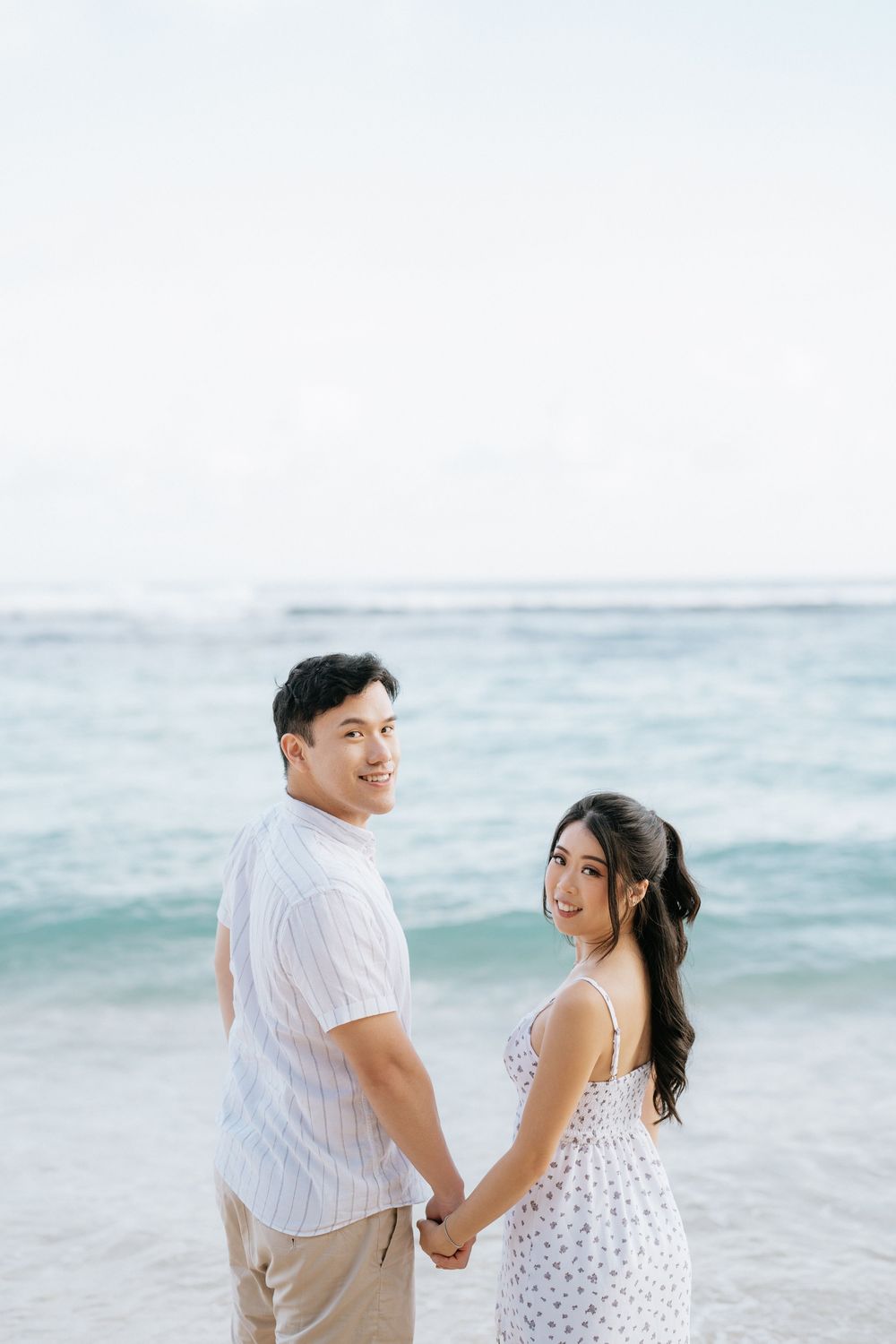 Romantic couple walking hand in hand along a serene beach with ocean waves in the background.