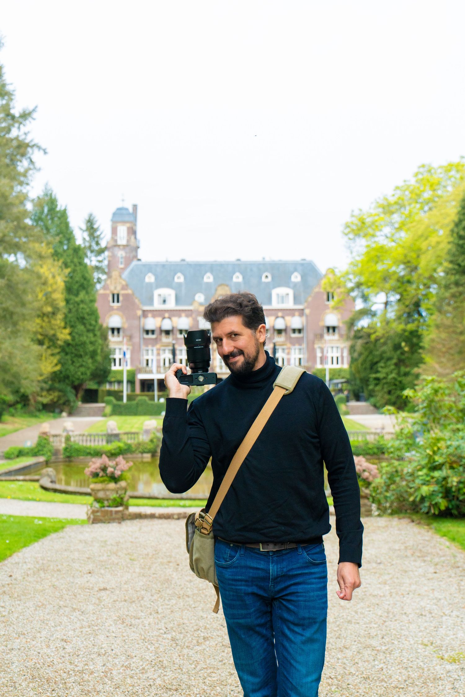Smiling man holding a camera and backpack stands in front of a grand European estate with a reflective pond.
