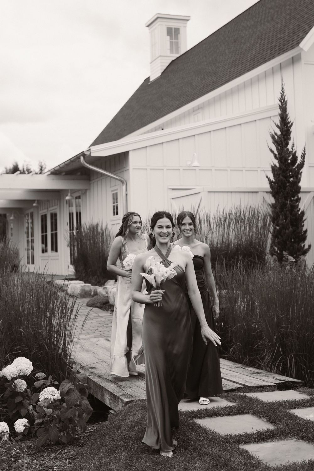Wedding party members share joyful moments outside a white barn building.