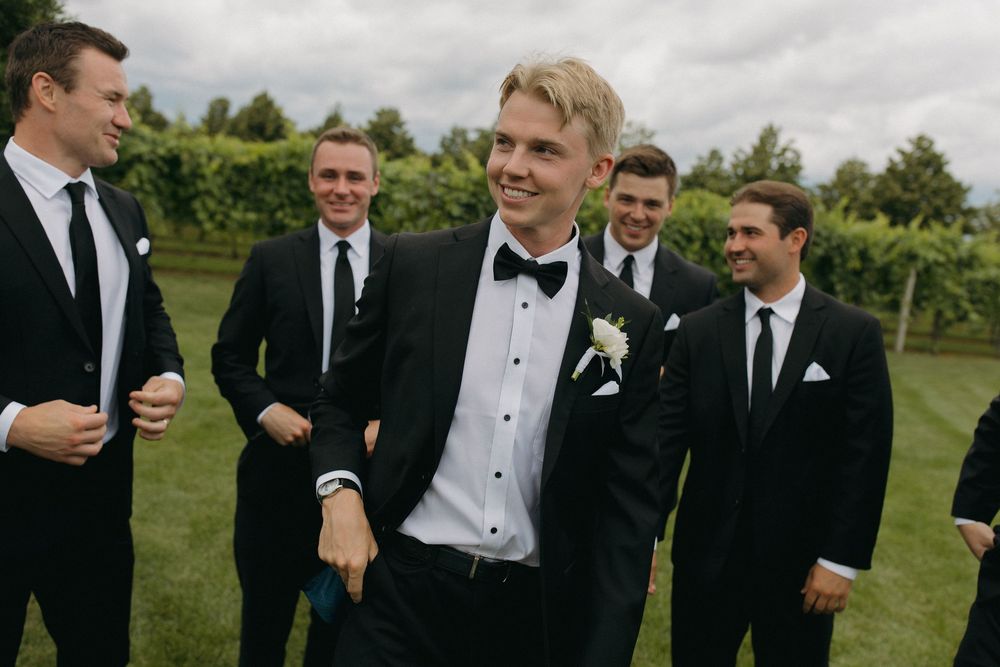 Groomsmen in black tuxedos and bowties smile together at an outdoor wedding venue.