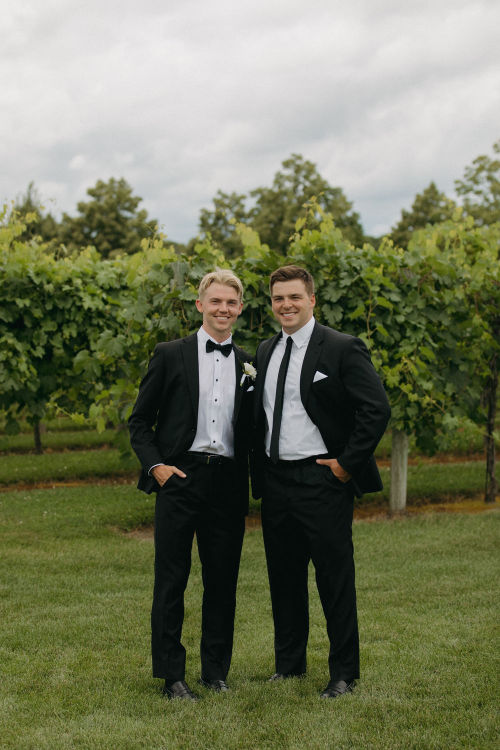 Groomsmen in formal attire share a candid moment in front of vineyard rows.