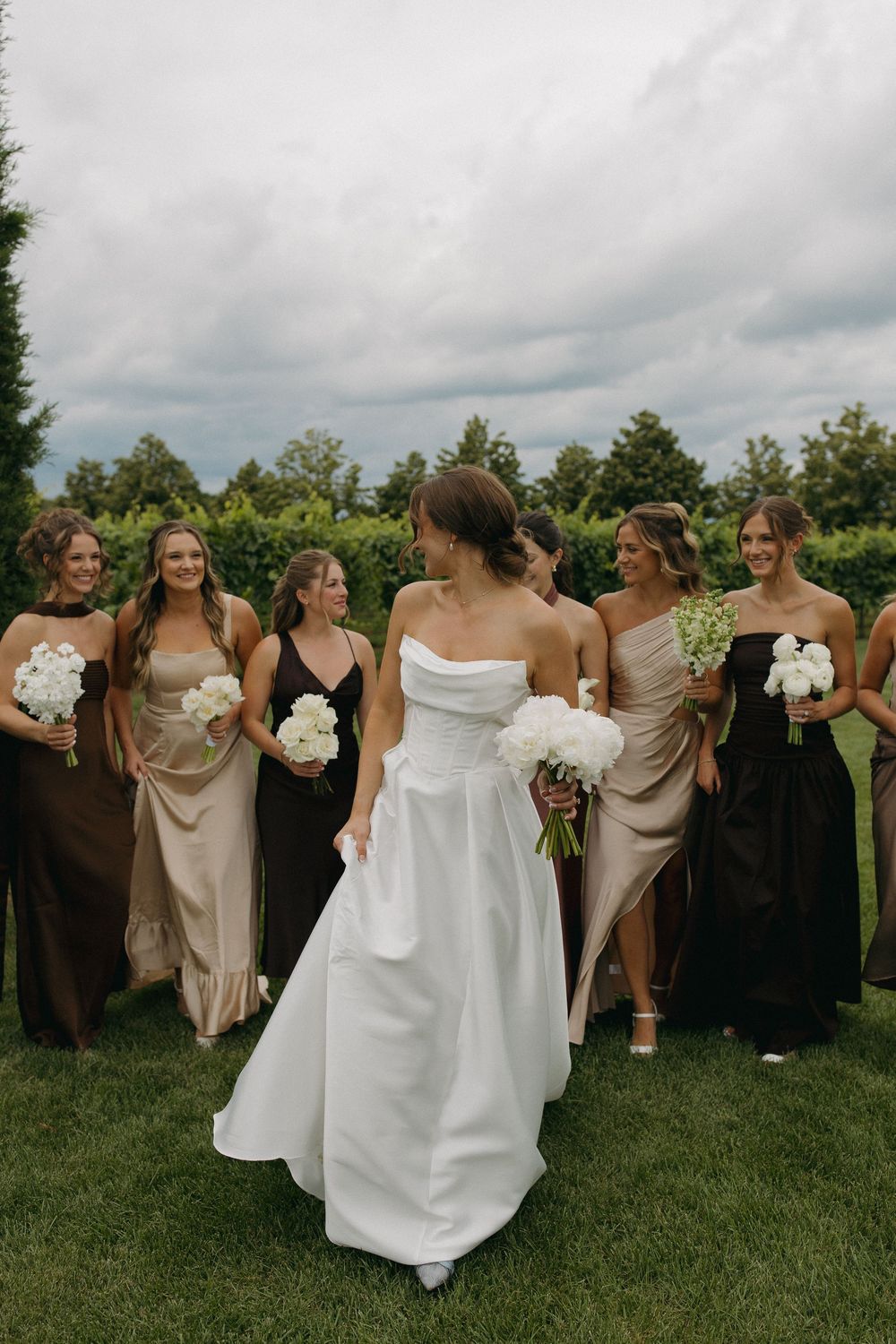 Wedding party poses together on a grassy lawn with cloudy skies in the background.