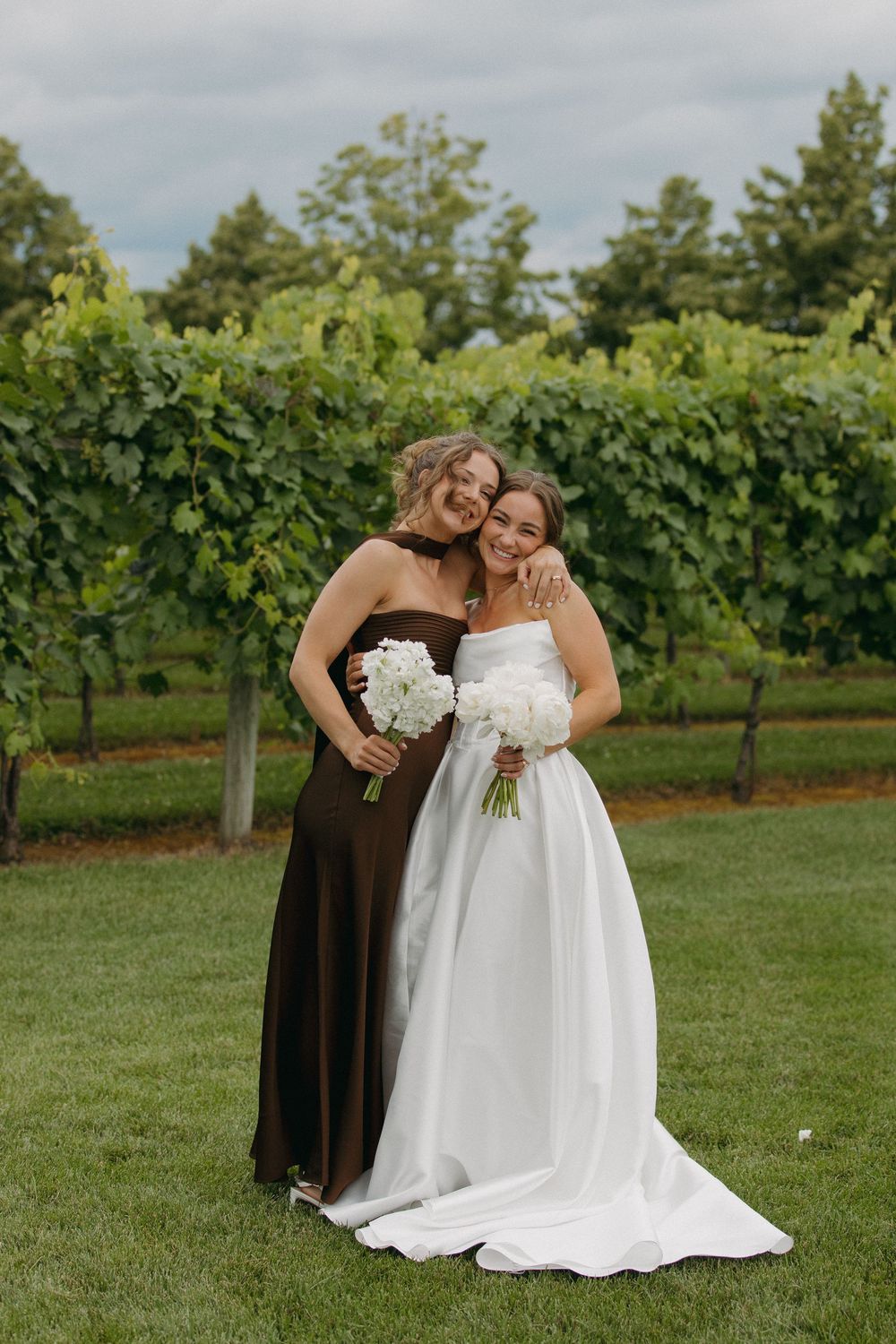 Wedding party members gather in a vineyard for photos wearing brown and white dresses.