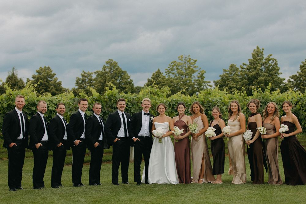 Large wedding party lines up for formal photos with bridesmaids in brown and groomsmen in black.