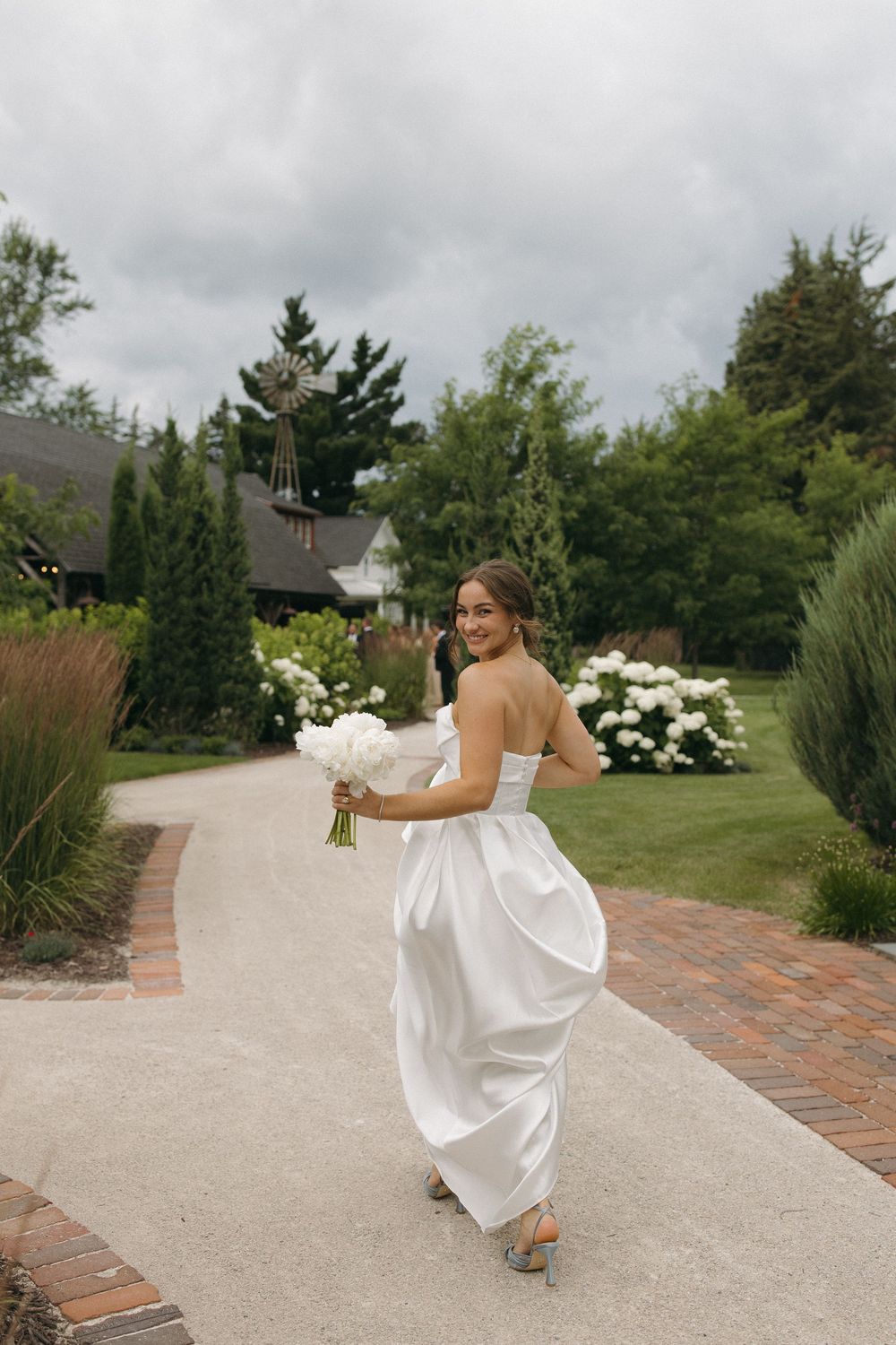 Person in white wedding dress walks playfully along a brick-lined path.