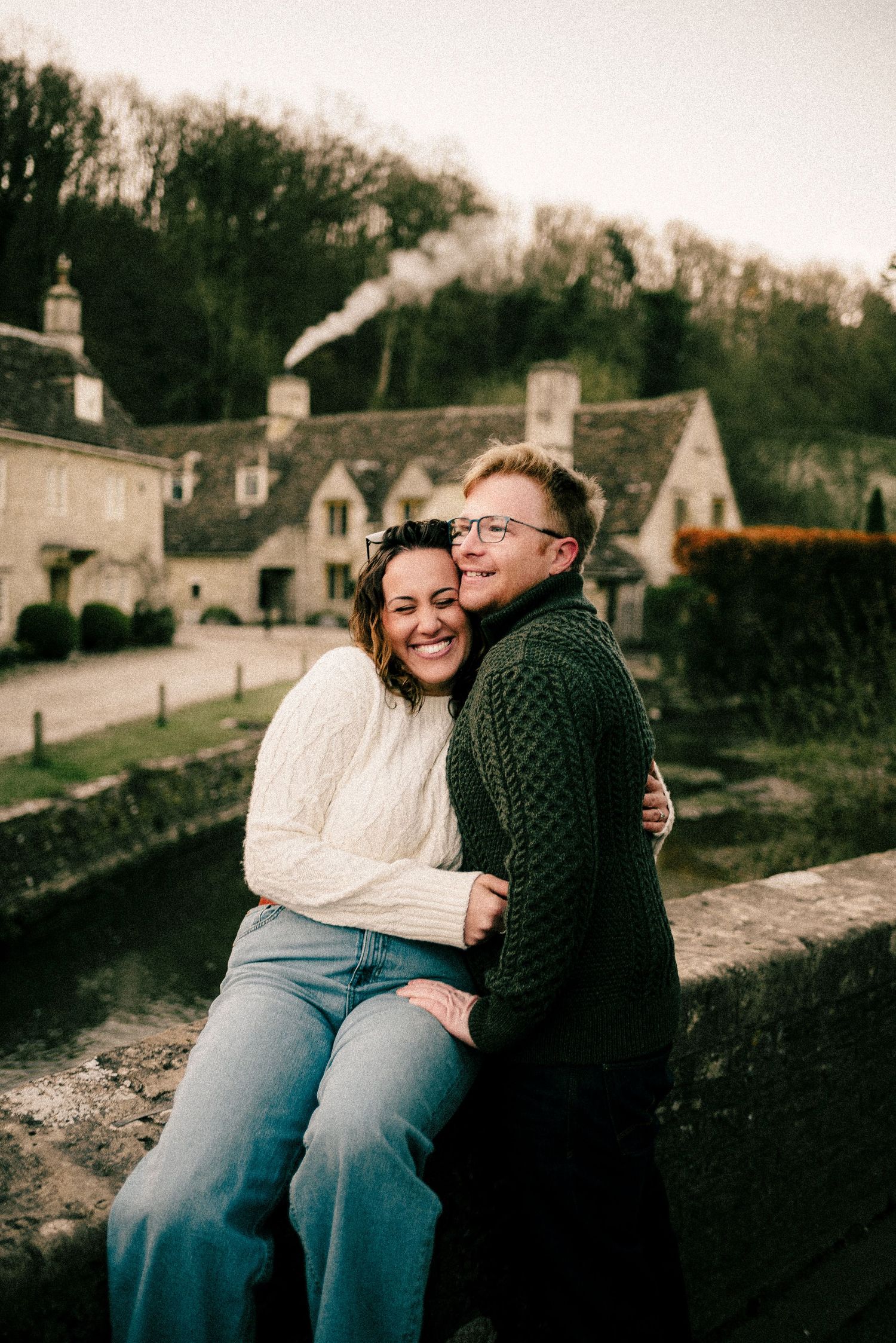 A couple sits close together sharing an embrace on a stone wall with rustic buildings visible in the background.