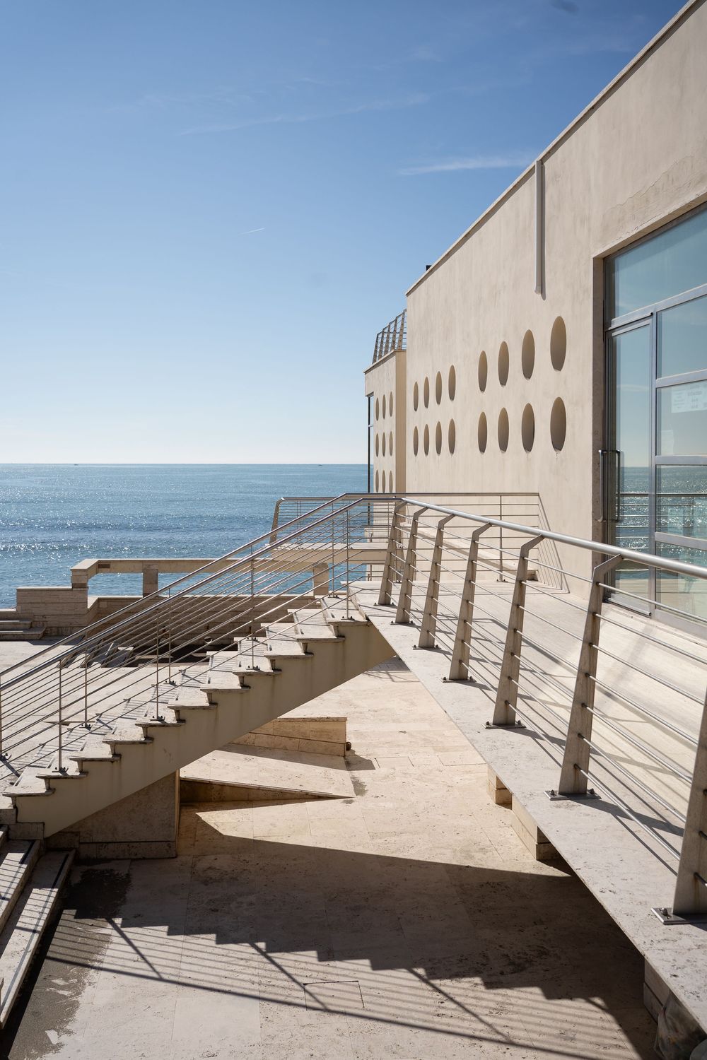 Modern oceanfront building with concrete staircase and metal railings overlooking the blue sea on a sunny day.