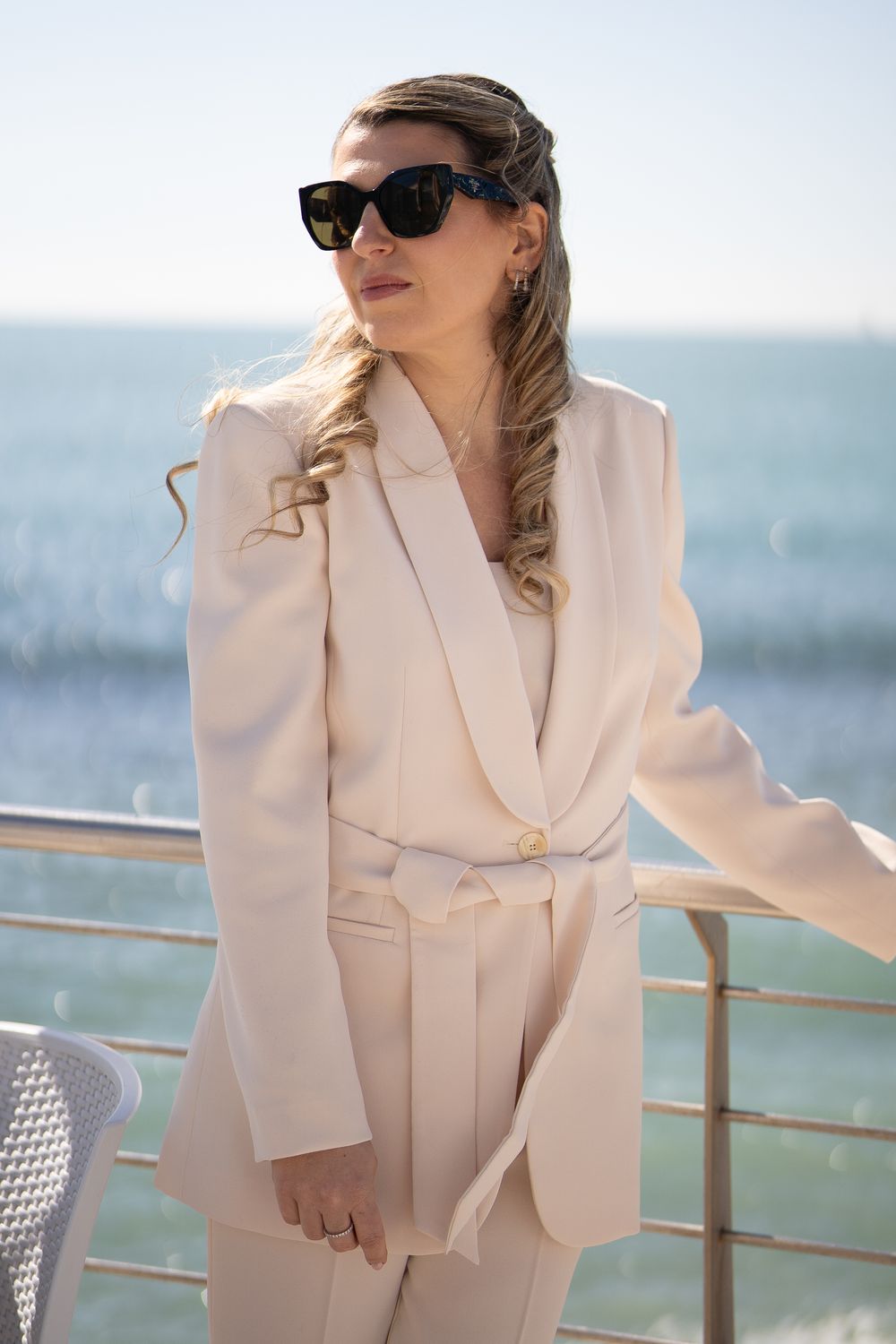 Person wearing white blazer and sunglasses sits overlooking ocean view from deck.