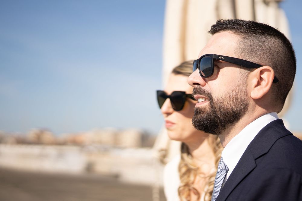 People in formal attire with sunglasses posing outdoors against bright sky.
