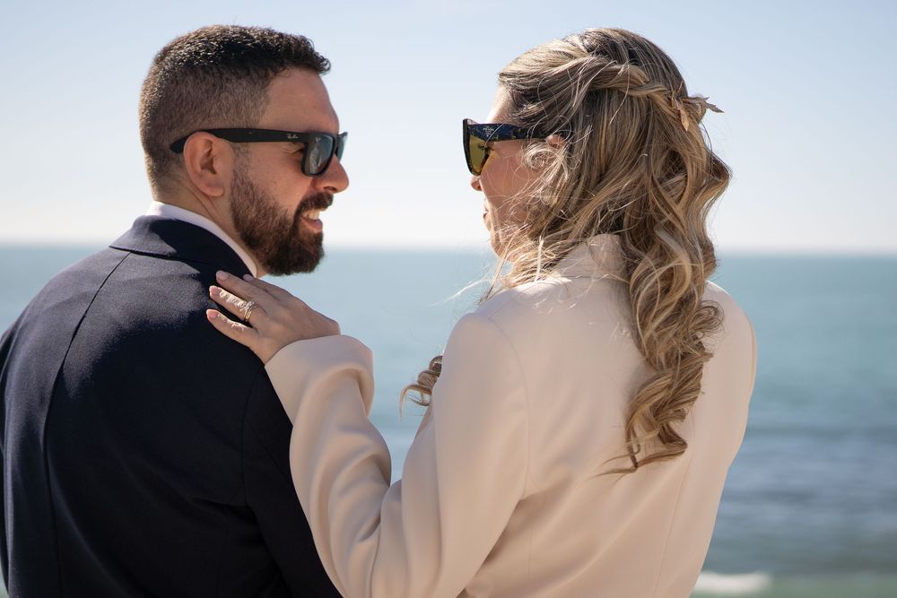 Romantic couple in formal attire embrace while looking at the ocean on a sunny day.