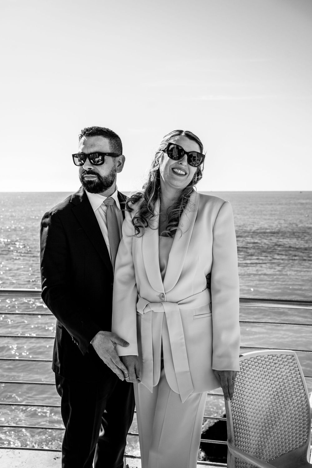 Stylish couple in contrasting black and white outfits pose together by the ocean railing.