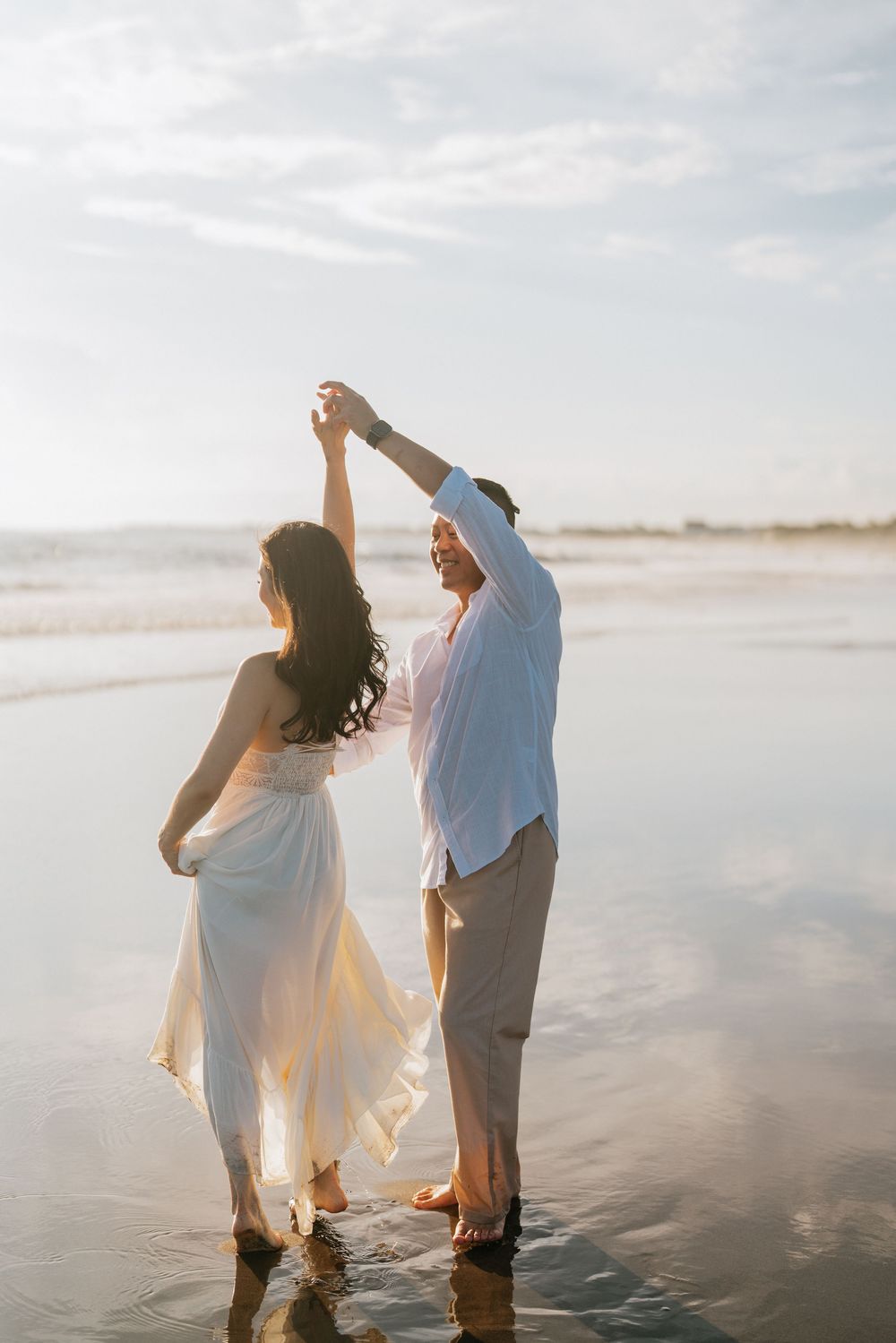 Couple dancing together on a serene beach during sunset with flowing white dress and casual attire.