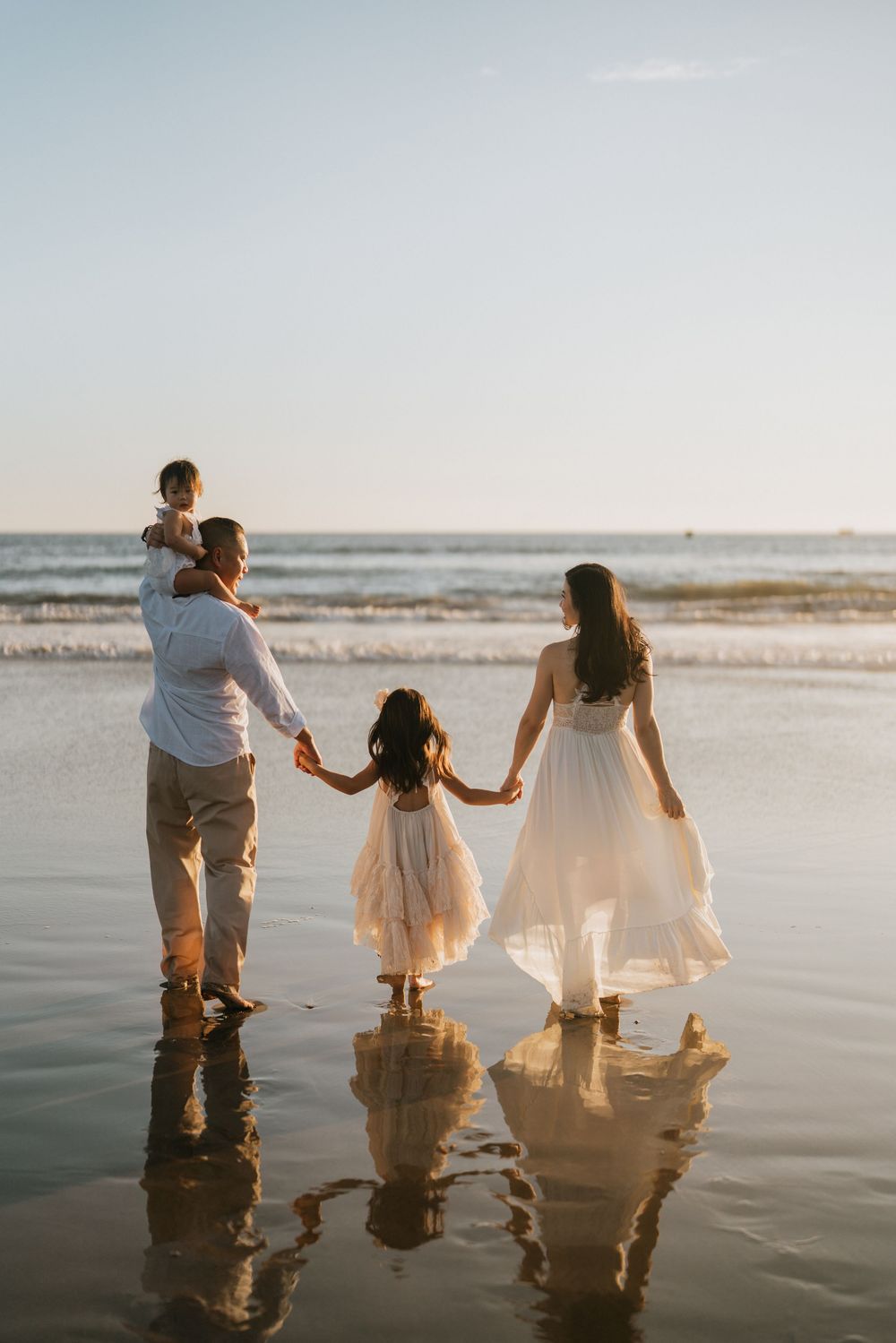 Group holding hands while walking in shallow ocean water at sunset creating reflections.
