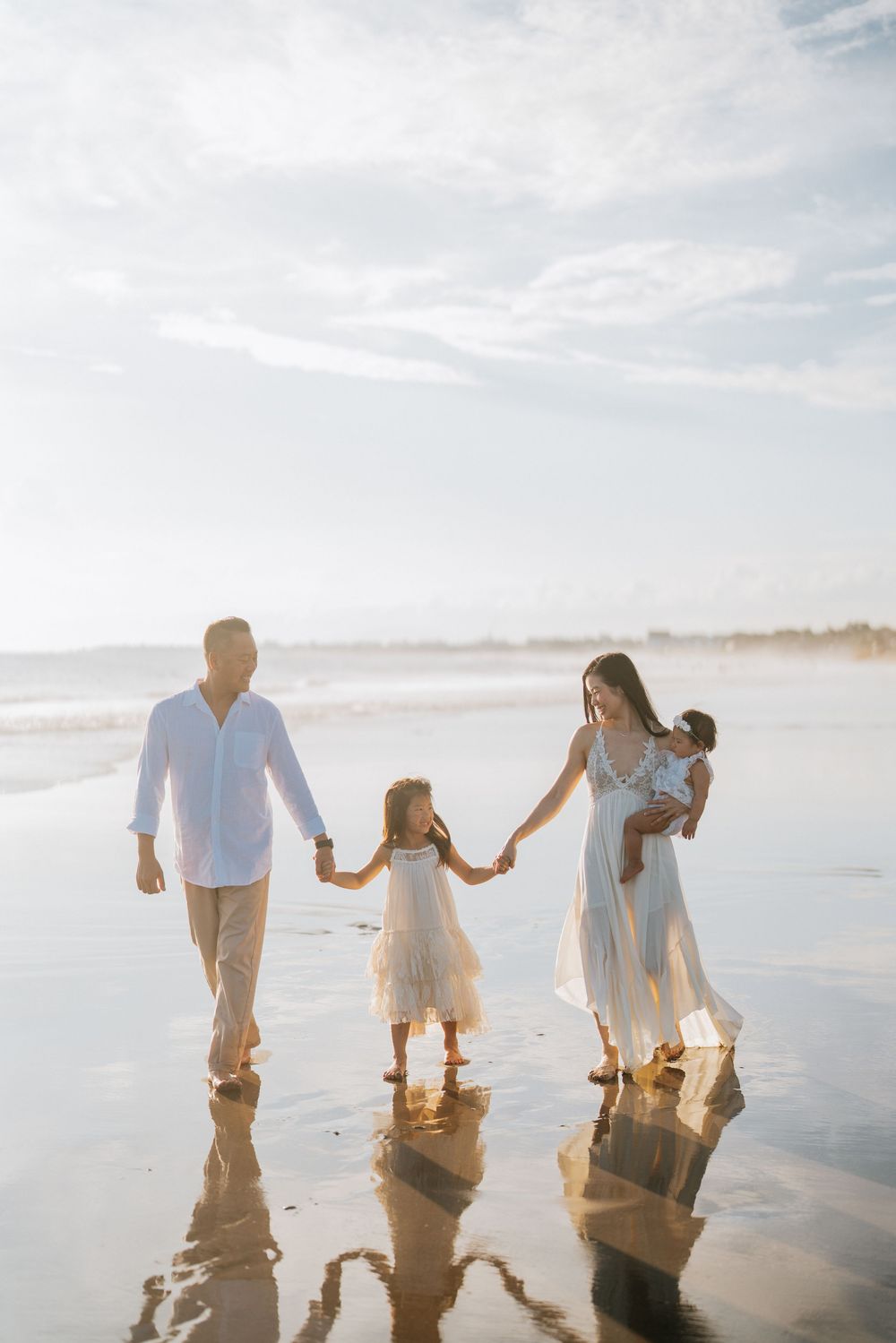 Family holding hands while walking on reflective wet beach sand at sunset.