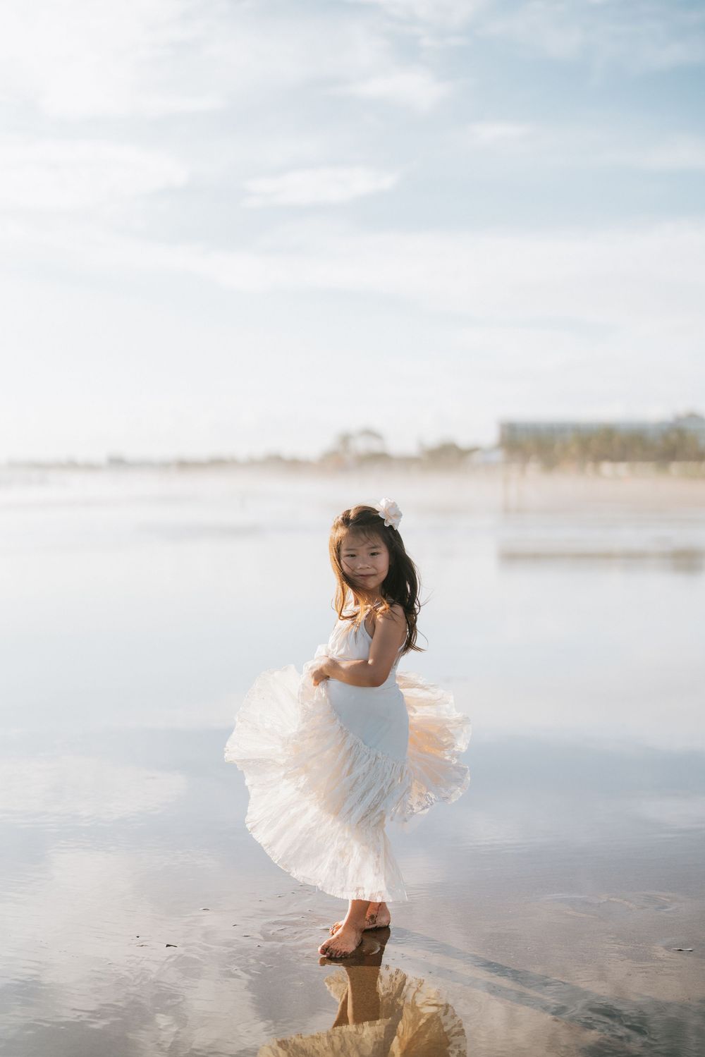 Young child in white dress twirling playfully on misty beach at sunrise.