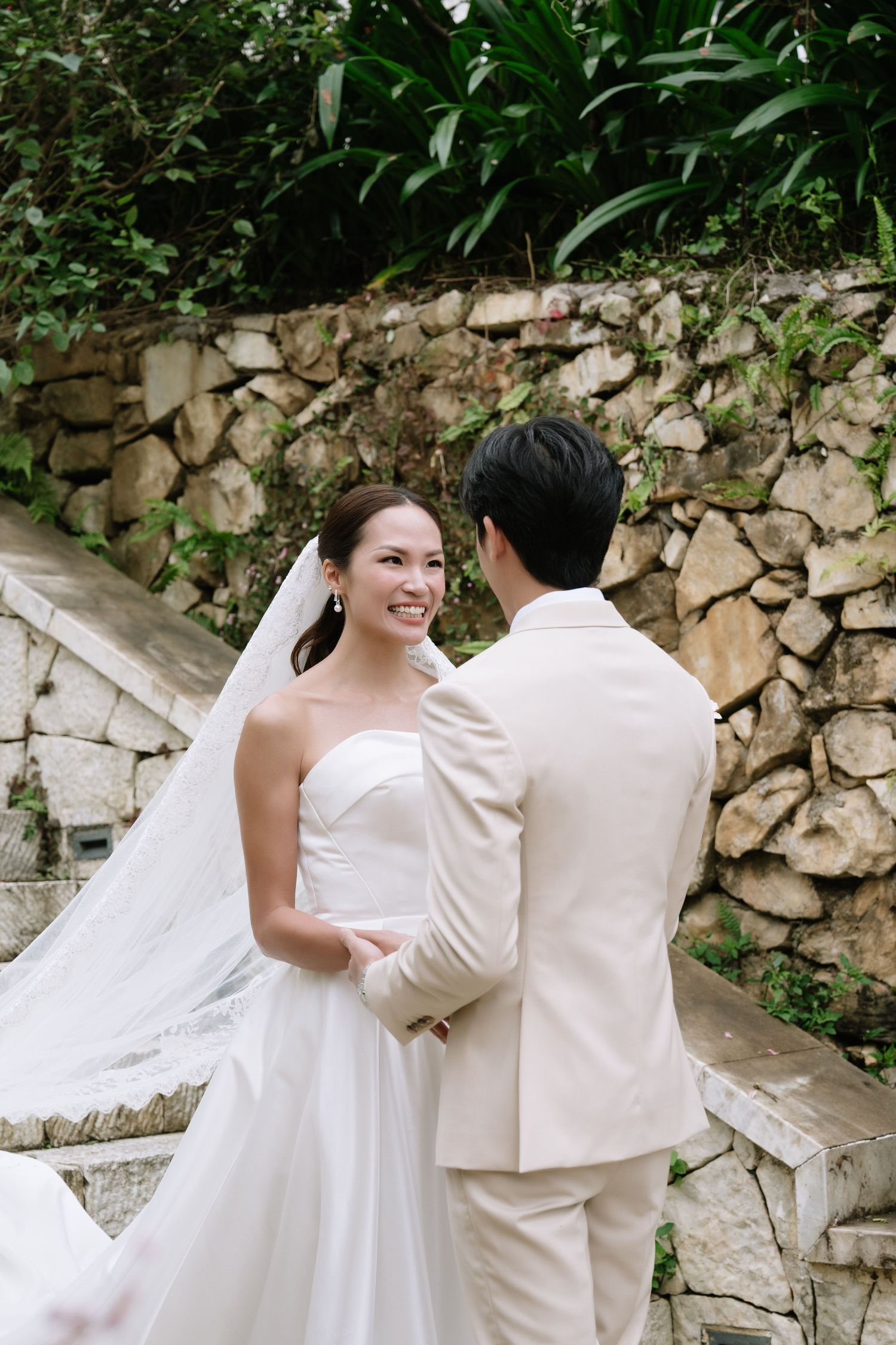 Smiling bride in white strapless gown holds grooms hands during outdoor first look on stone steps.