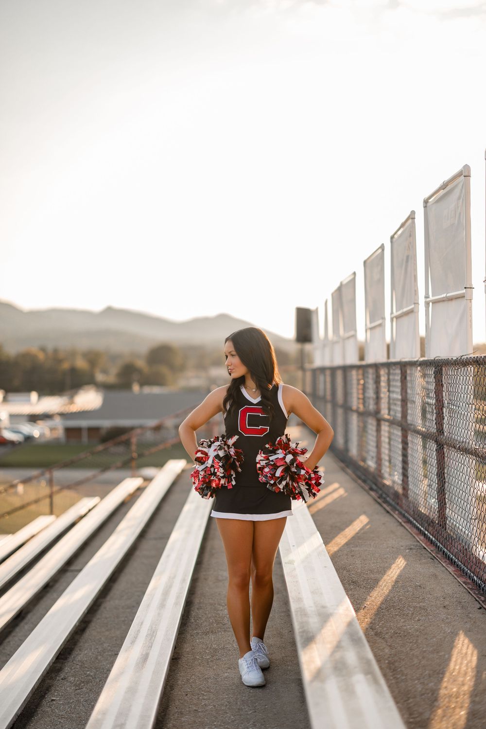Cheerleader in black uniform and pom poms sits on bleachers during golden hour with scenic mountain backdrop.