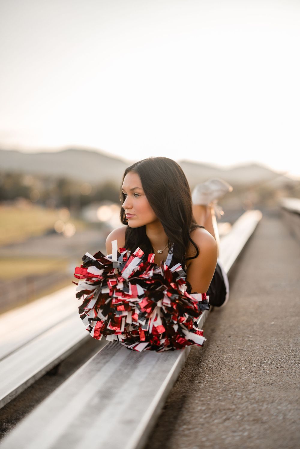 A cheerleader with red and white pom poms poses on the sidelines of a stadium at sunset.