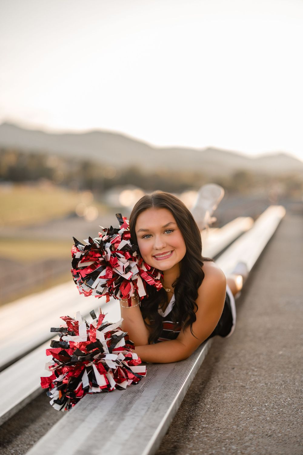 A cheerleader poses with red, black and white pom-poms on a runway while laying down in outdoor photoshoot.