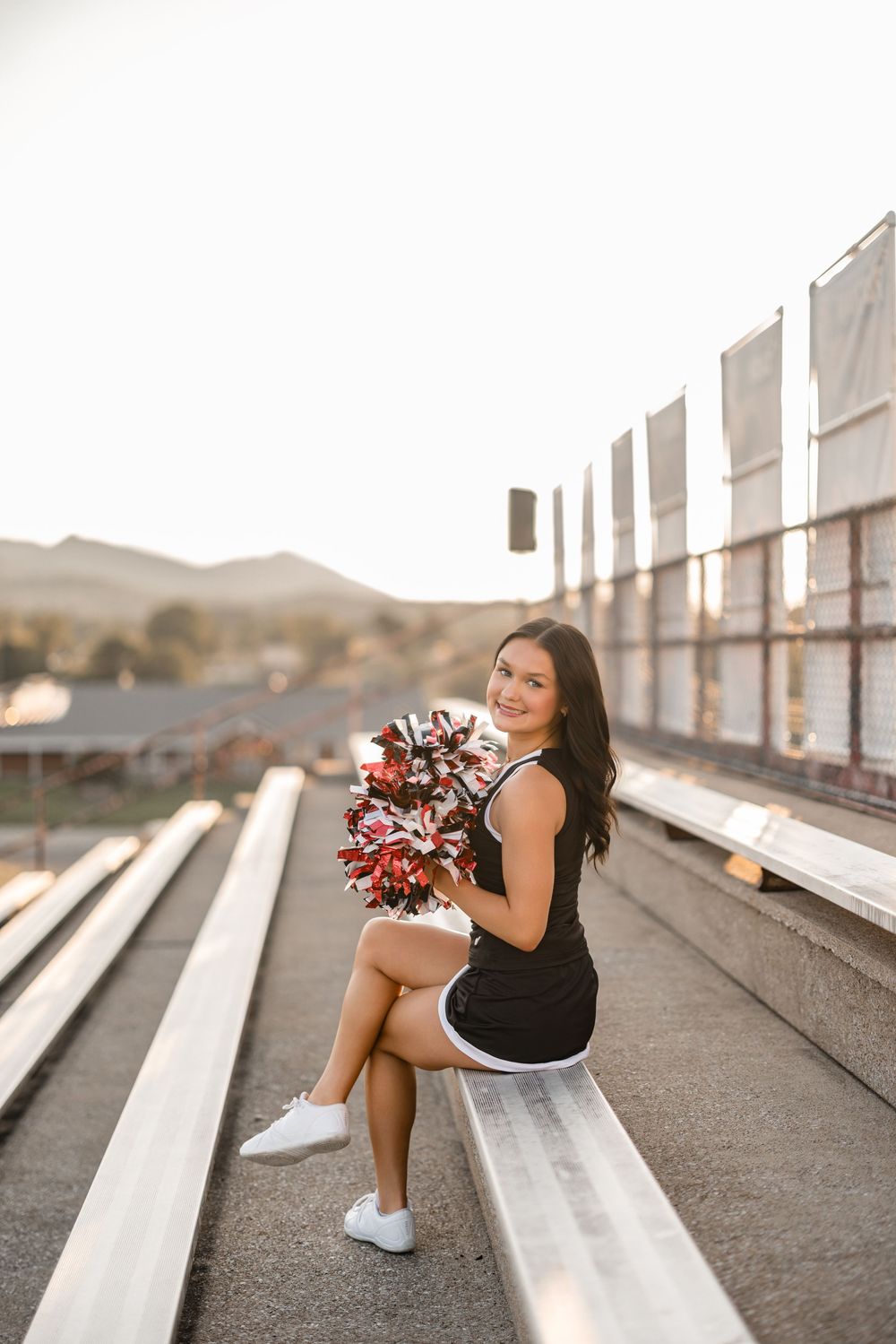 Cheerleader in black uniform sits casually on stadium bleachers during sunset photoshoot.