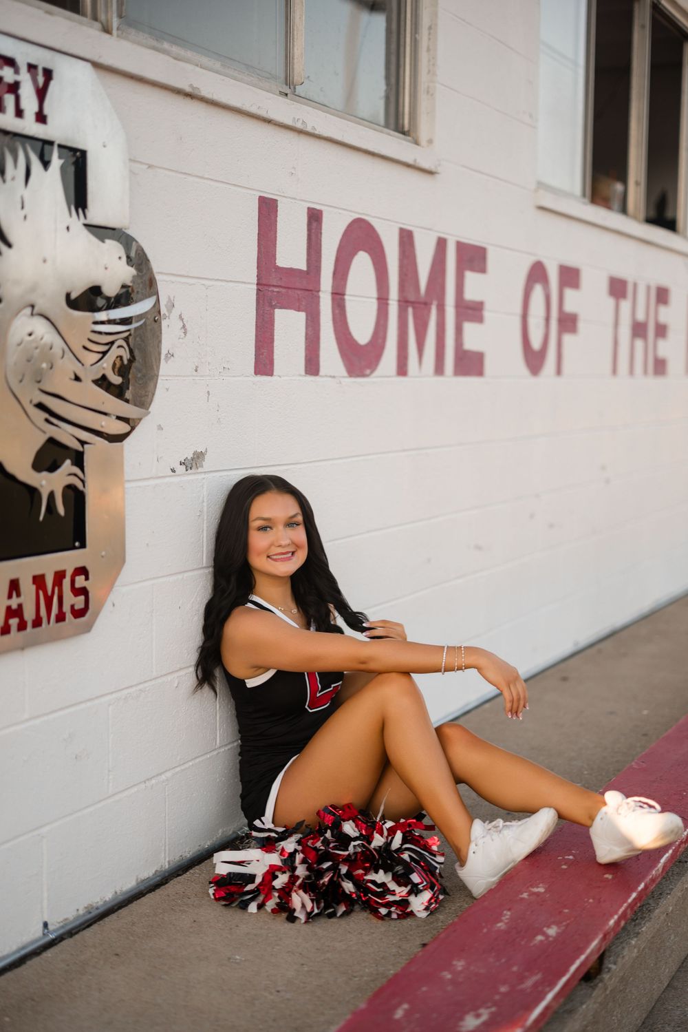 Cheerleader posing casually against white wall with 'Home of the' text in school hallway.