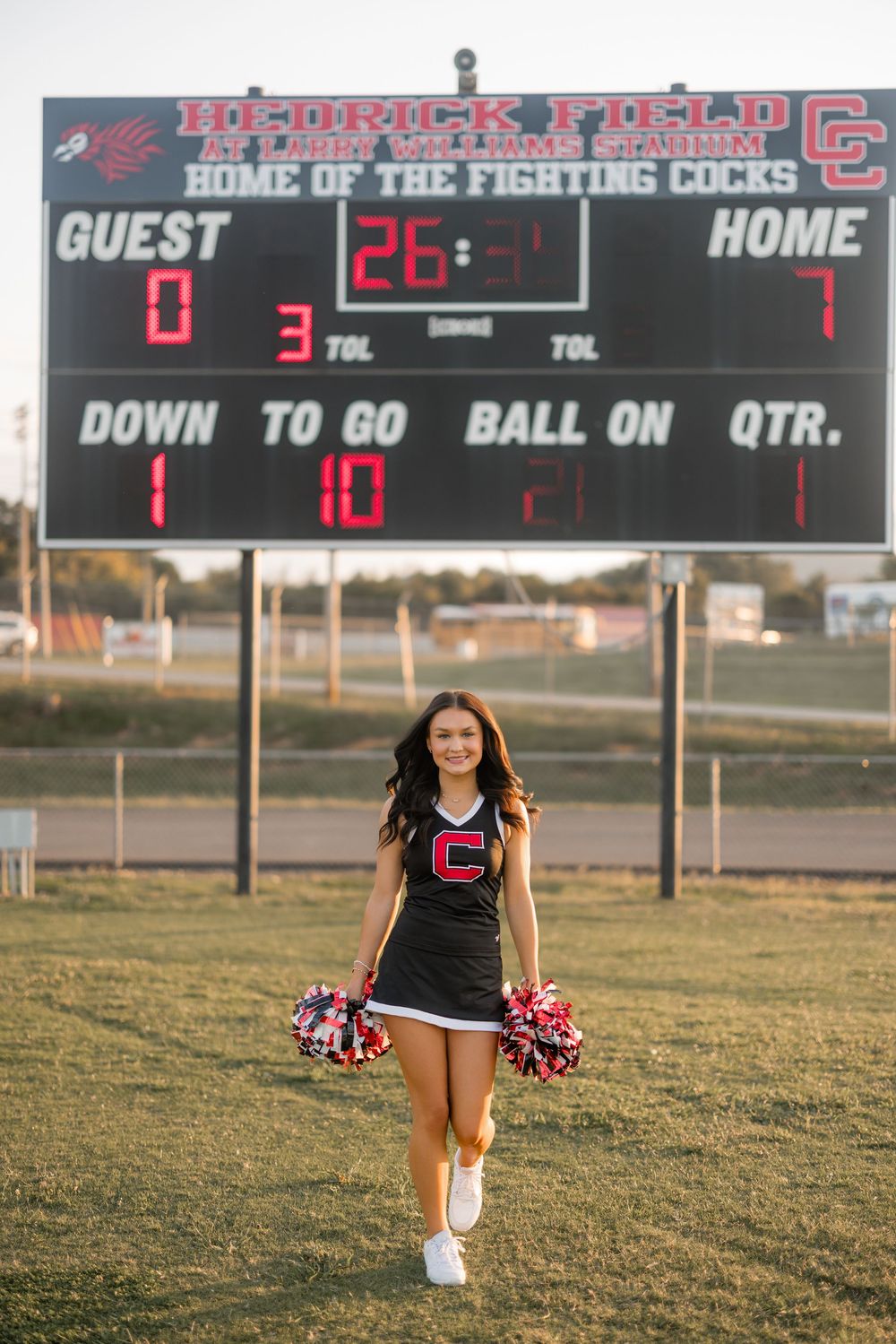 A cheerleader performs on a football field at sunset with scoreboards visible in the background.