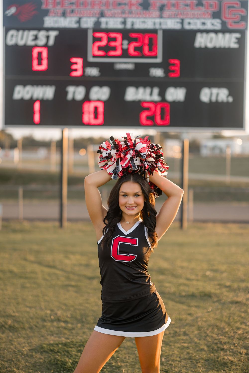 Cheerleader performing on sports field with scoreboard and stadium lights in background at sunset.