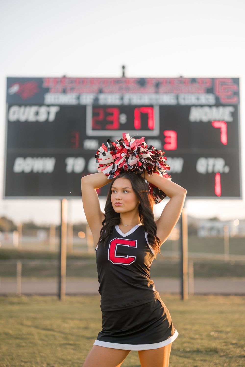 A cheerleader in a black uniform with 'C' letter holds pom-poms overhead in front of a scoreboard at sunset.