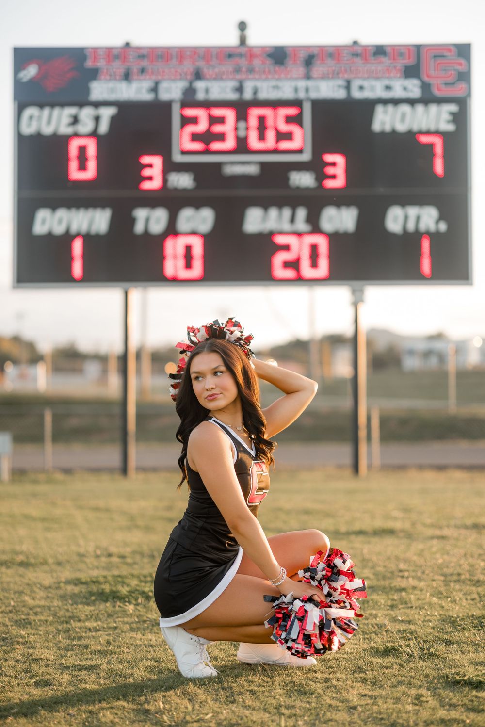 A cheerleader poses with pom poms under a scoreboard at sunset on an athletic field.