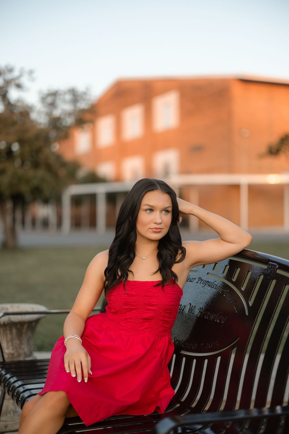 A woman in a red dress sits elegantly on a park bench at golden hour.
