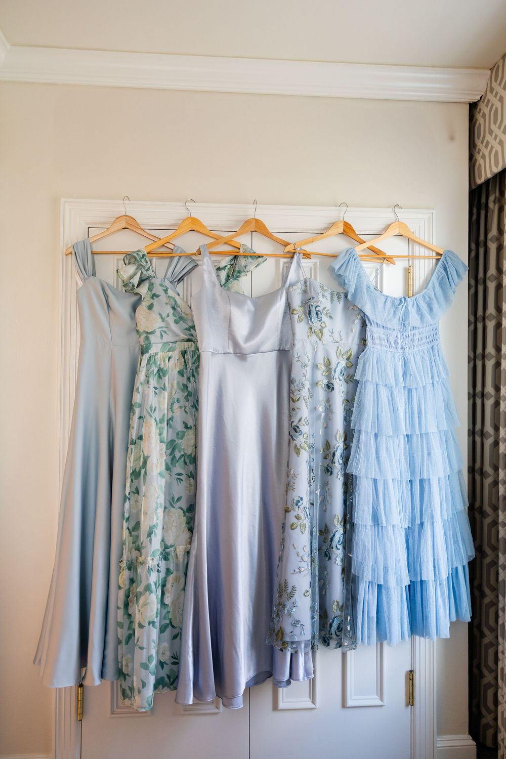 Light blue and floral bridesmaid dresses hanging on wooden hangers against a white wall.
