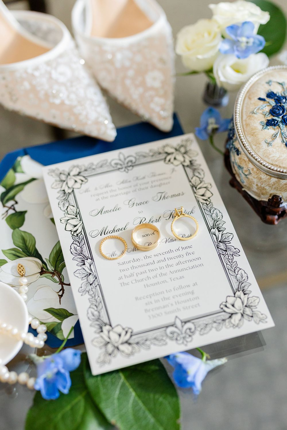 Wedding invitations with ornate frames and gold rings displayed with blue flowers.