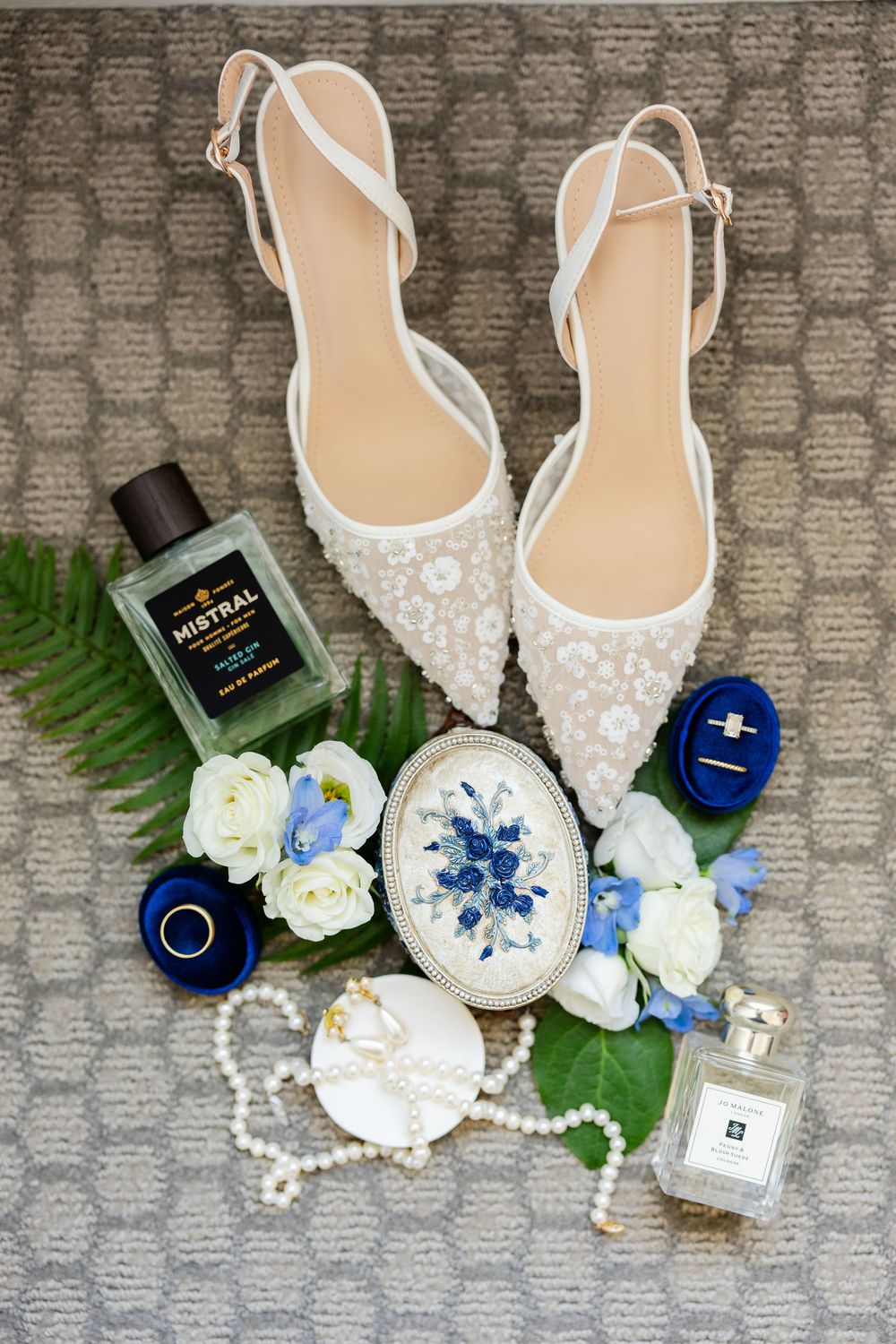 Wedding accessories including white beaded shoes, perfume bottles, rings, pearls and blue and white flowers arranged on grey carpet.