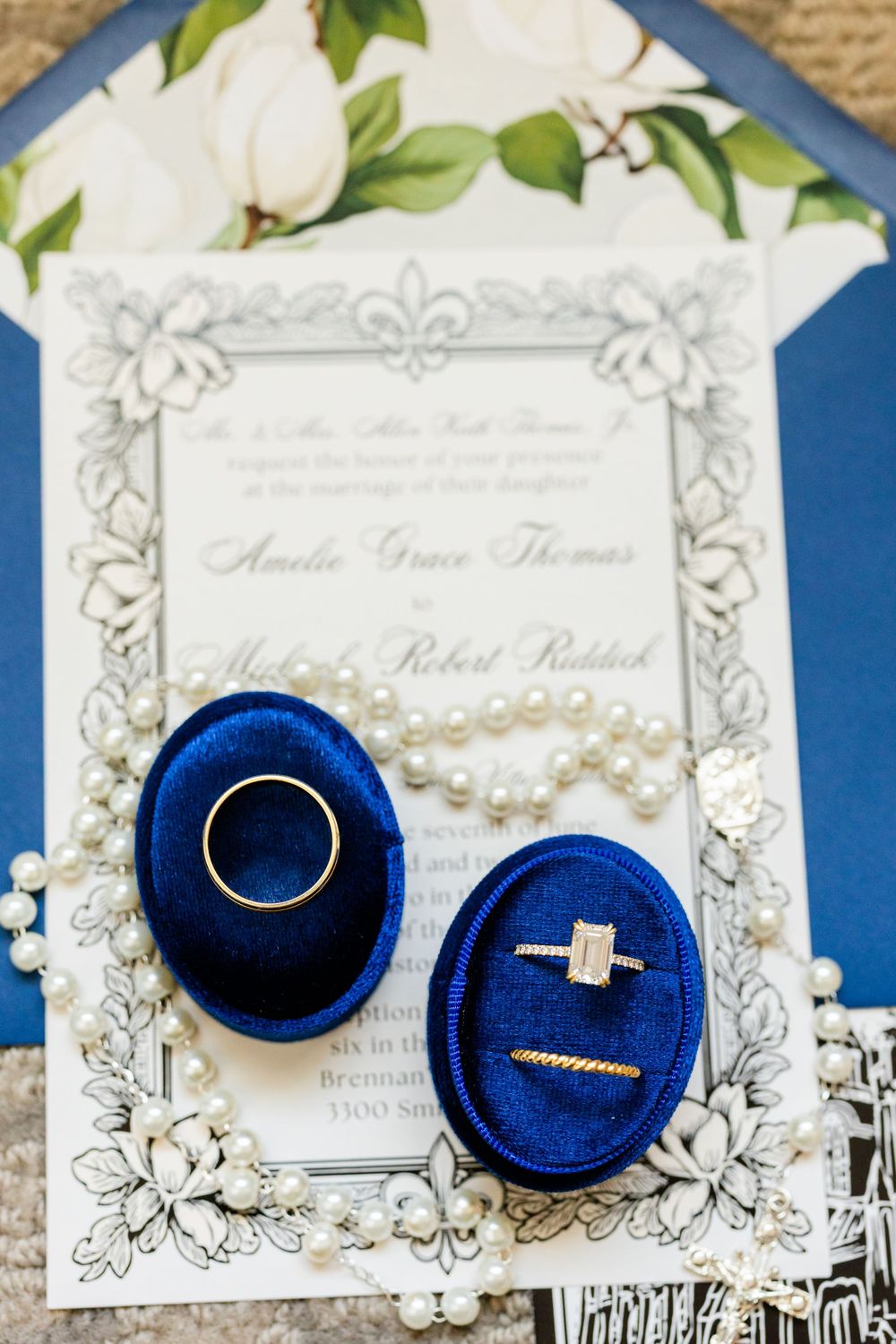 Wedding rings displayed in elegant navy blue velvet ring boxes alongside an ornate invitation on white paper.