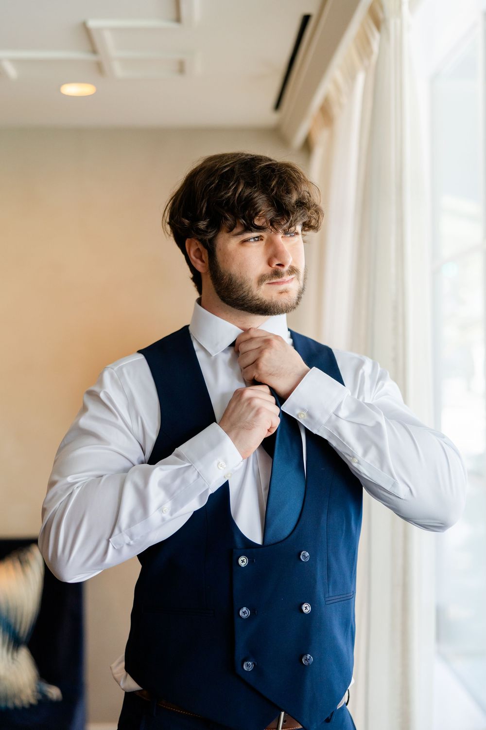 A person in a navy vest and white shirt adjusts their tie while standing by a window.