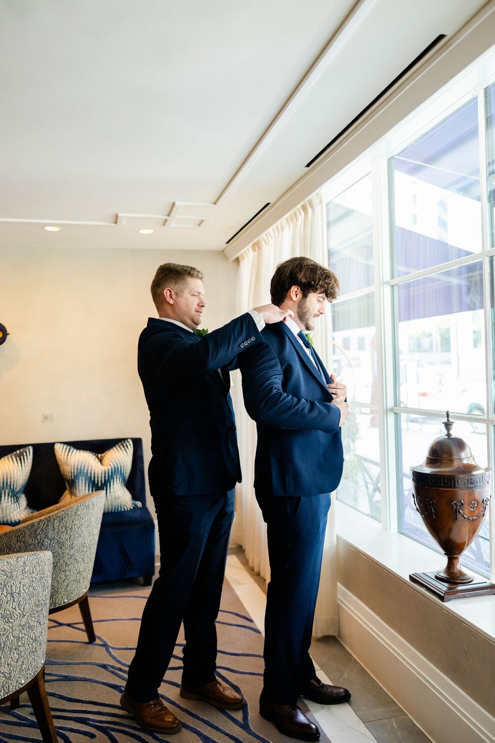 Two people in navy suits standing by a window as one helps adjust the other's jacket.