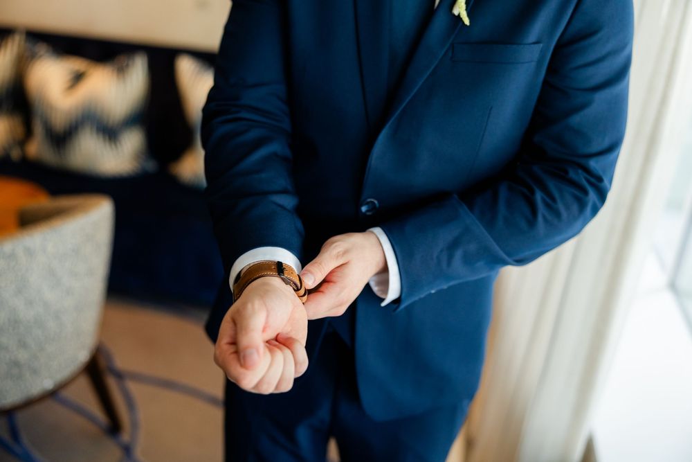 Well-dressed person in a navy blue suit adjusts their cufflink before an event while standing in natural light.