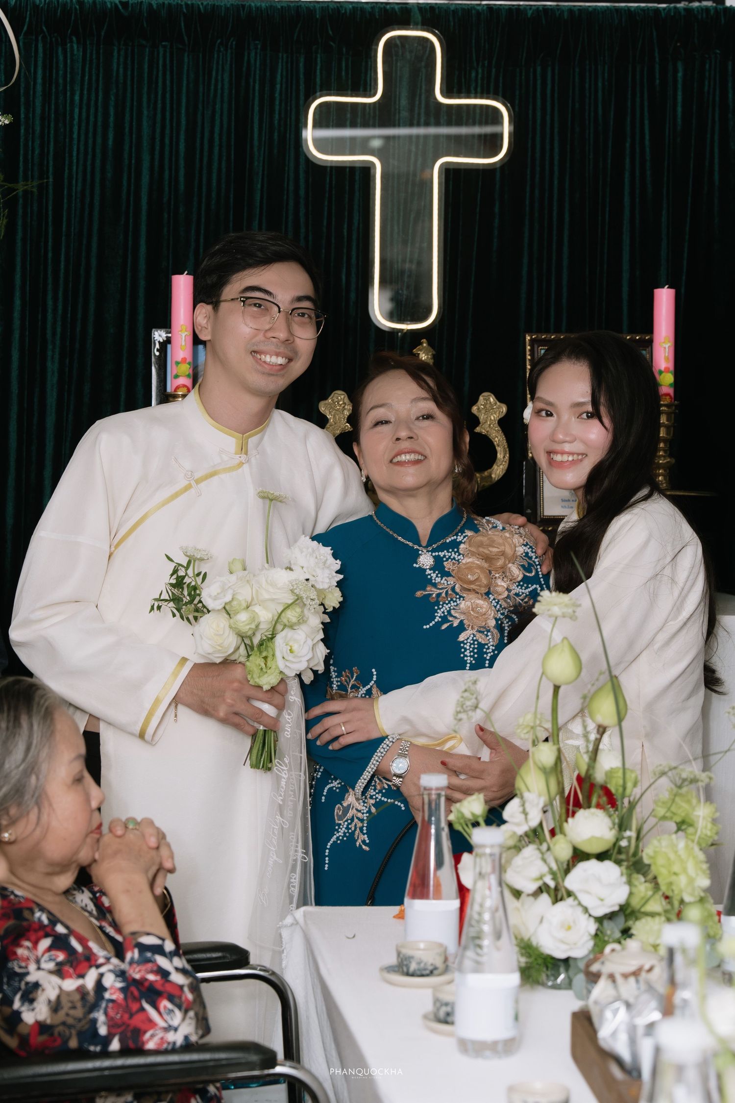 Bride and groom in white ao dai pose with woman in blue ao dai at Vietnamese Christian wedding ceremony altar.