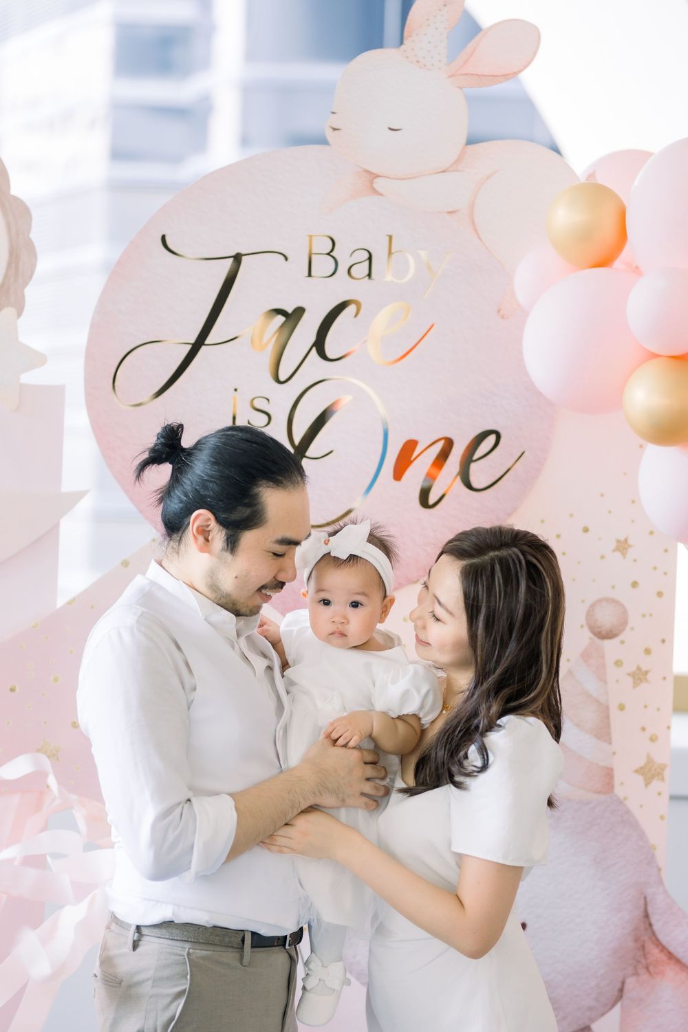 Family embraces and laughs together during celebration with pink balloon decorations and gold lettering on wall.