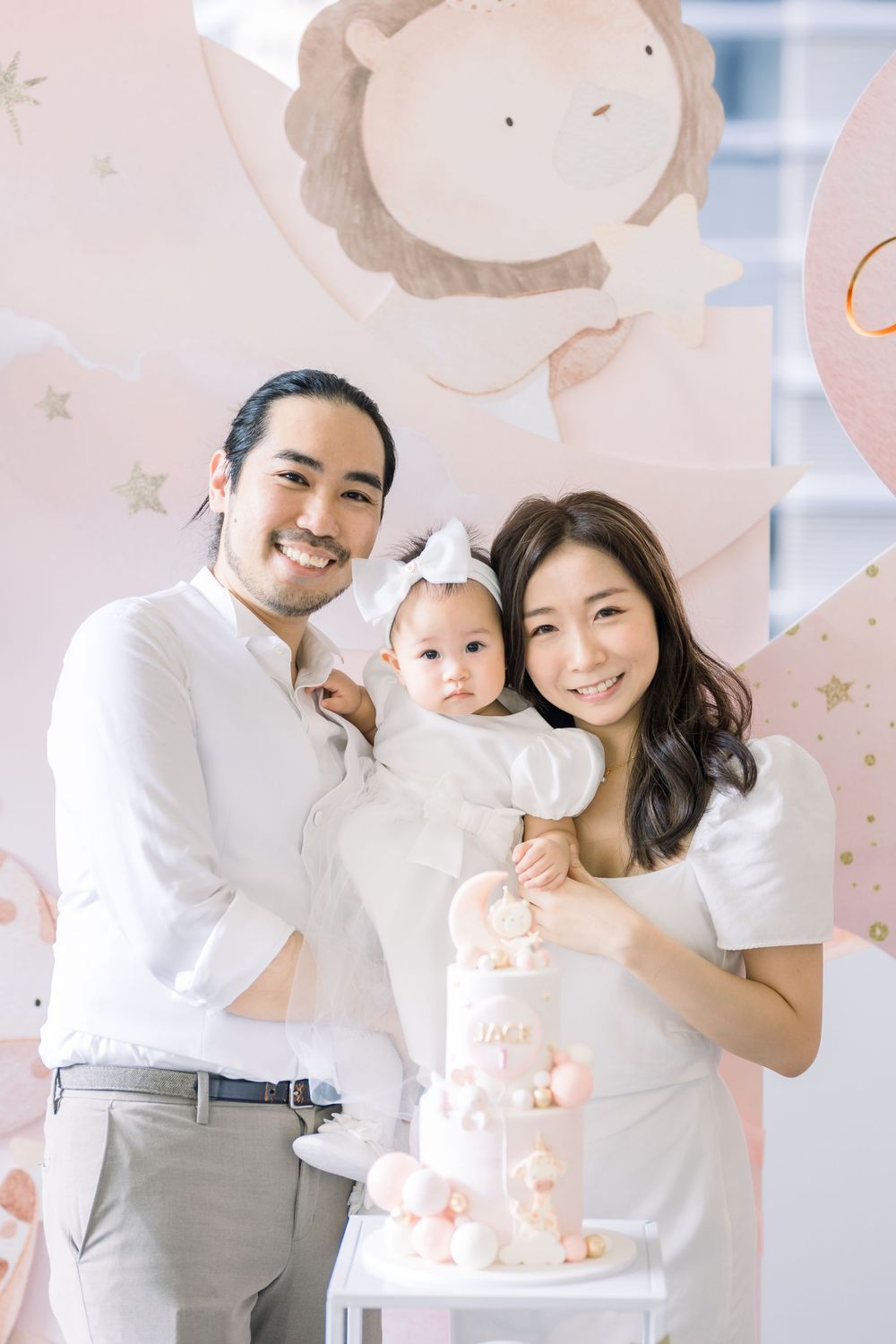 Family celebrates special occasion with elegant pink and white cake against pastel balloon-decorated backdrop.