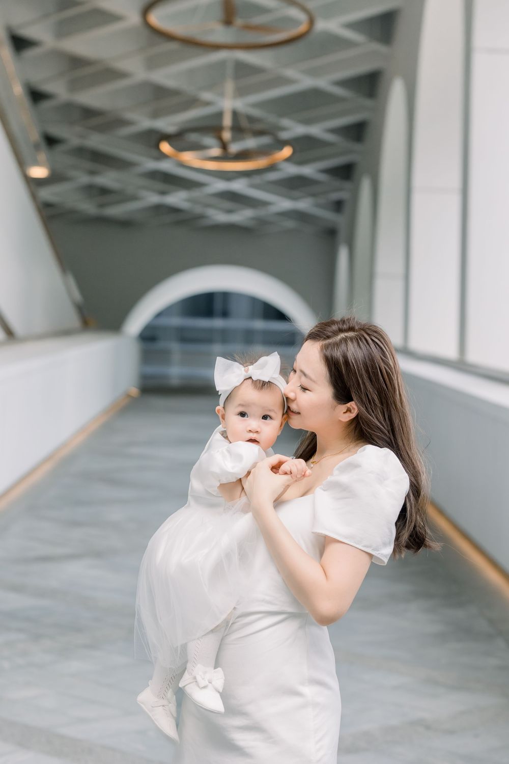 A woman in a white dress holds a baby wearing a white outfit and bow in an elegant hallway with modern pendant lights.