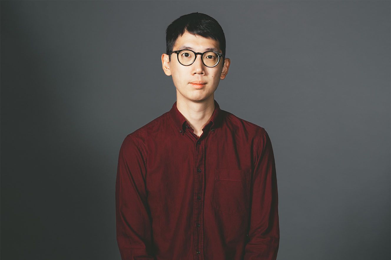 Person in burgundy sweater and round glasses sits against gray background in professional portrait series.