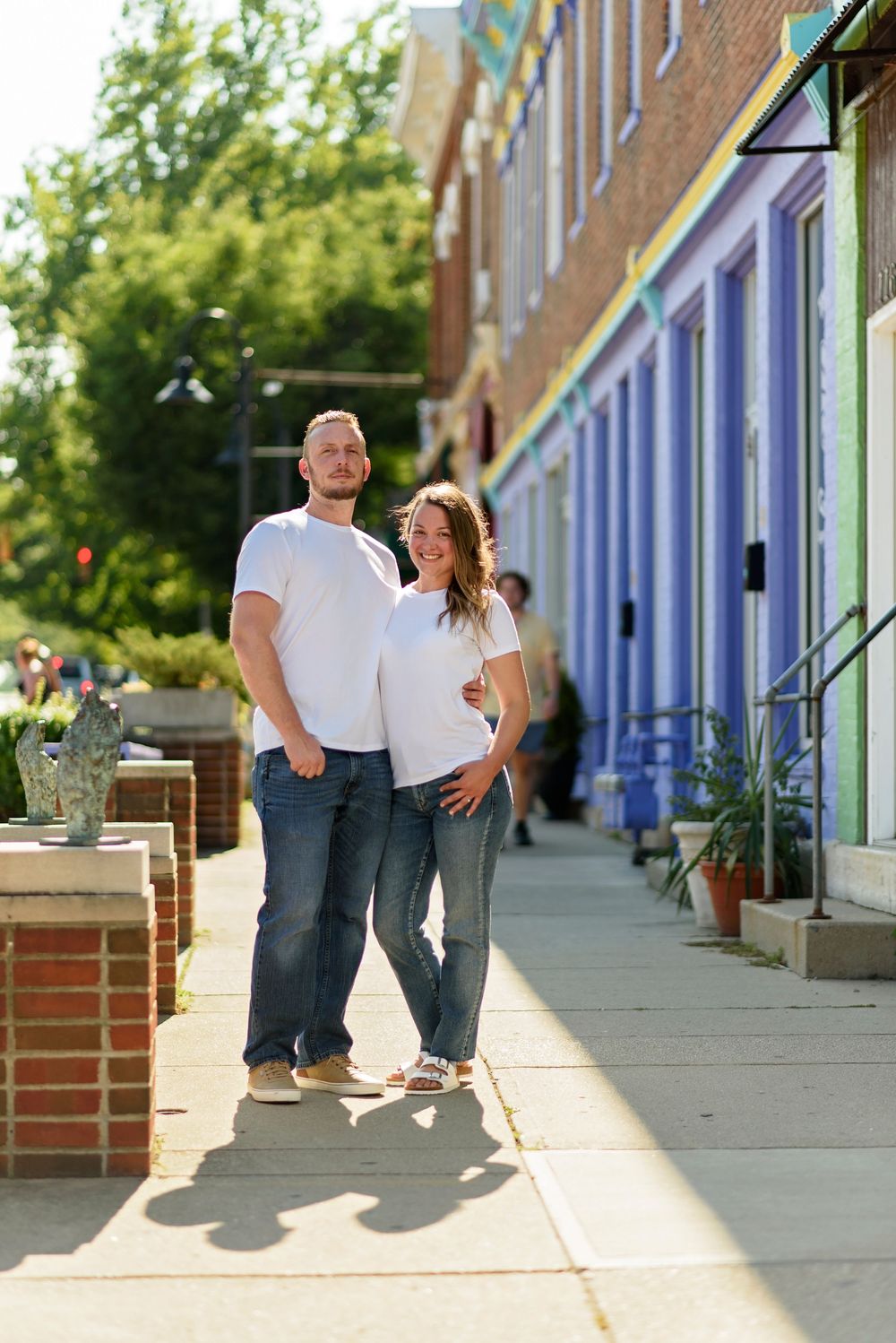 Couple poses casually against colorful storefront walls on sunny sidewalk.