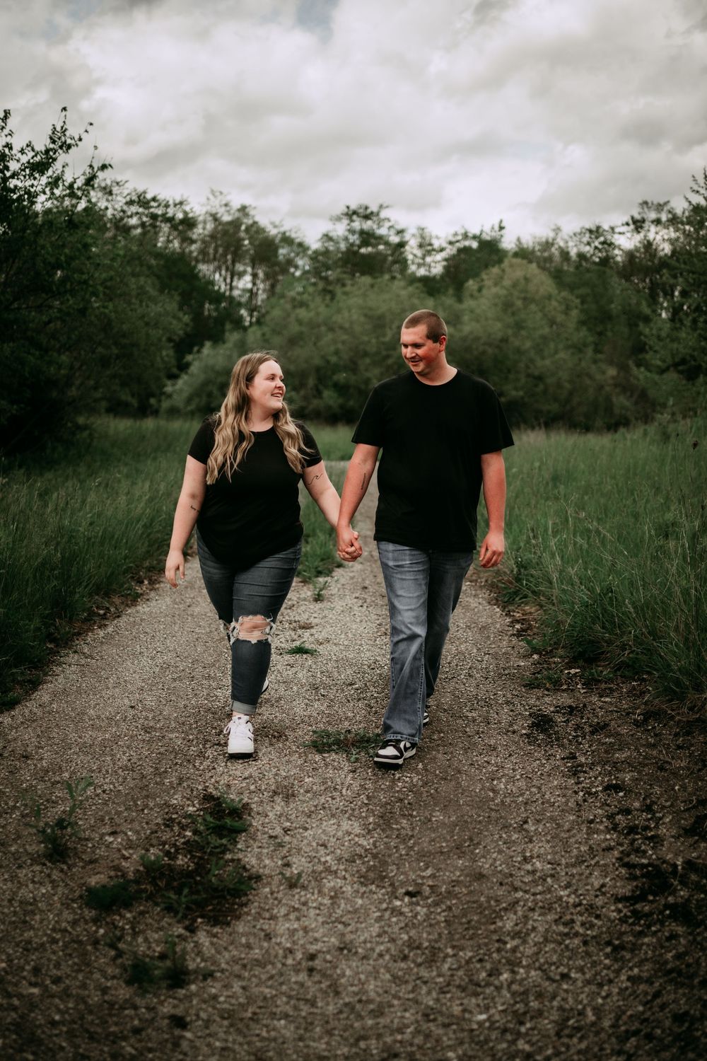 Couple walking together on a gravel path surrounded by lush green trees and grass on a cloudy day.